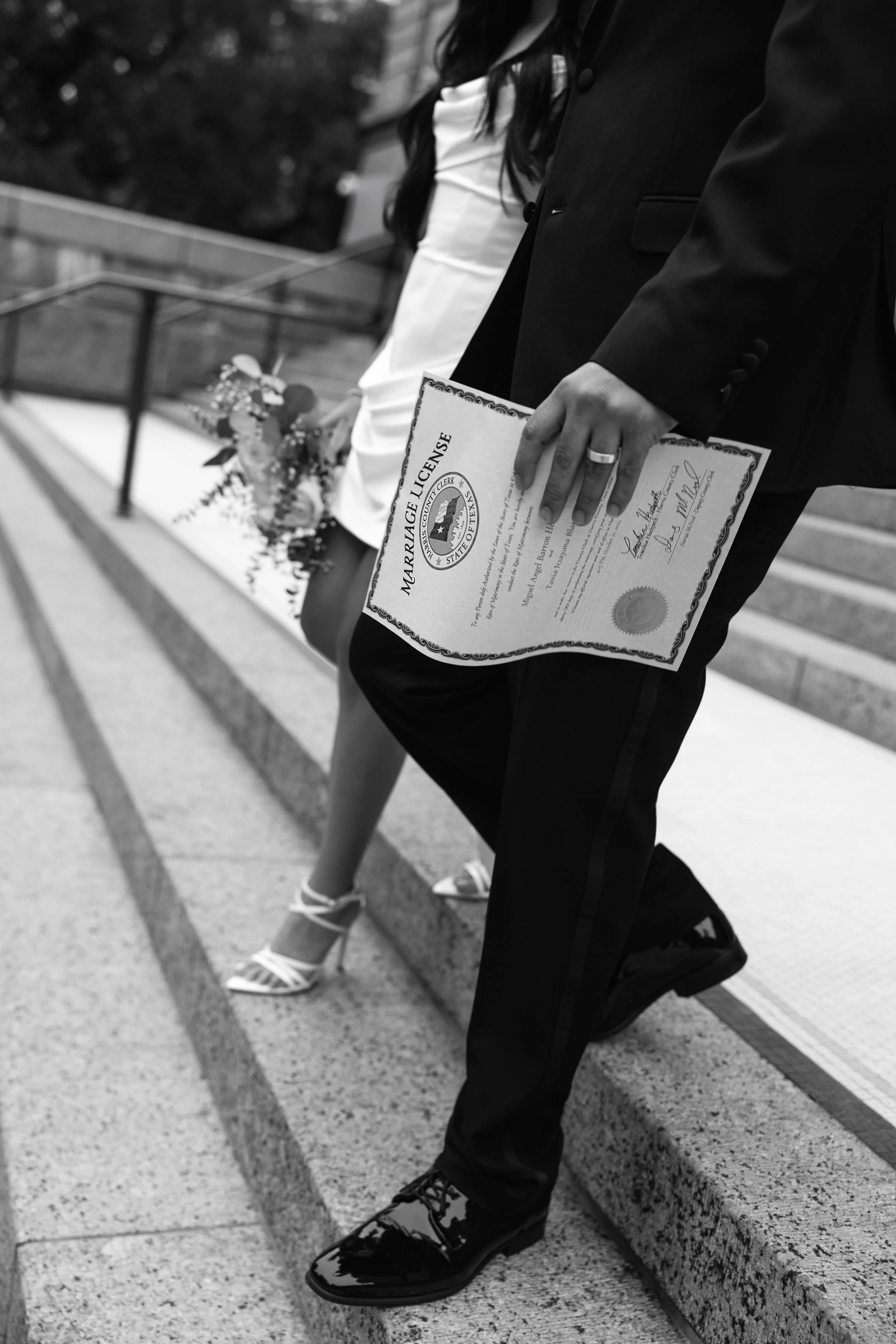 A person holding a marriage license in a black suit at a wedding ceremony, with a woman in a white dress and heels standing nearby.