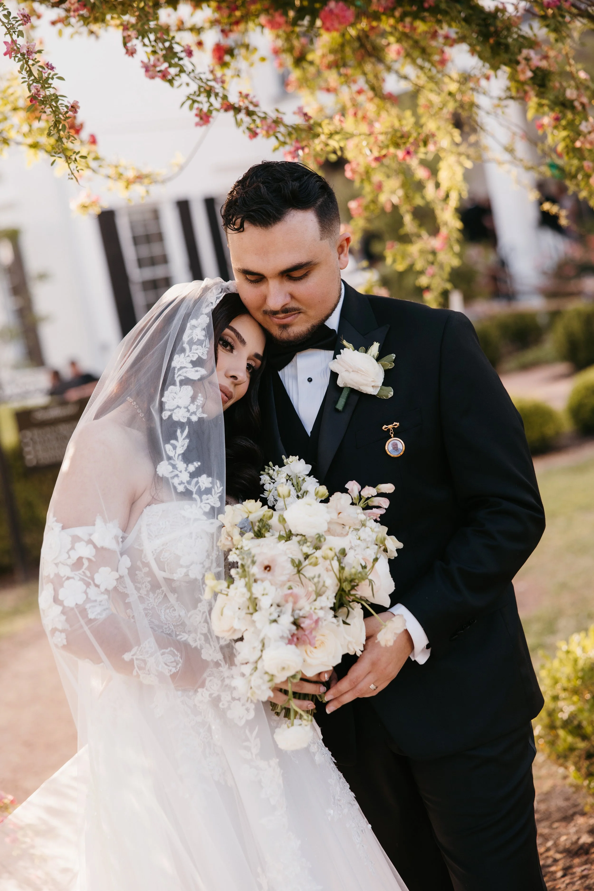 A bride and groom embrace outdoors during their wedding, with pink blossoms in the background. The bride is wearing a white lace wedding gown and veil, and the groom is in a black tuxedo with a white boutonniere.