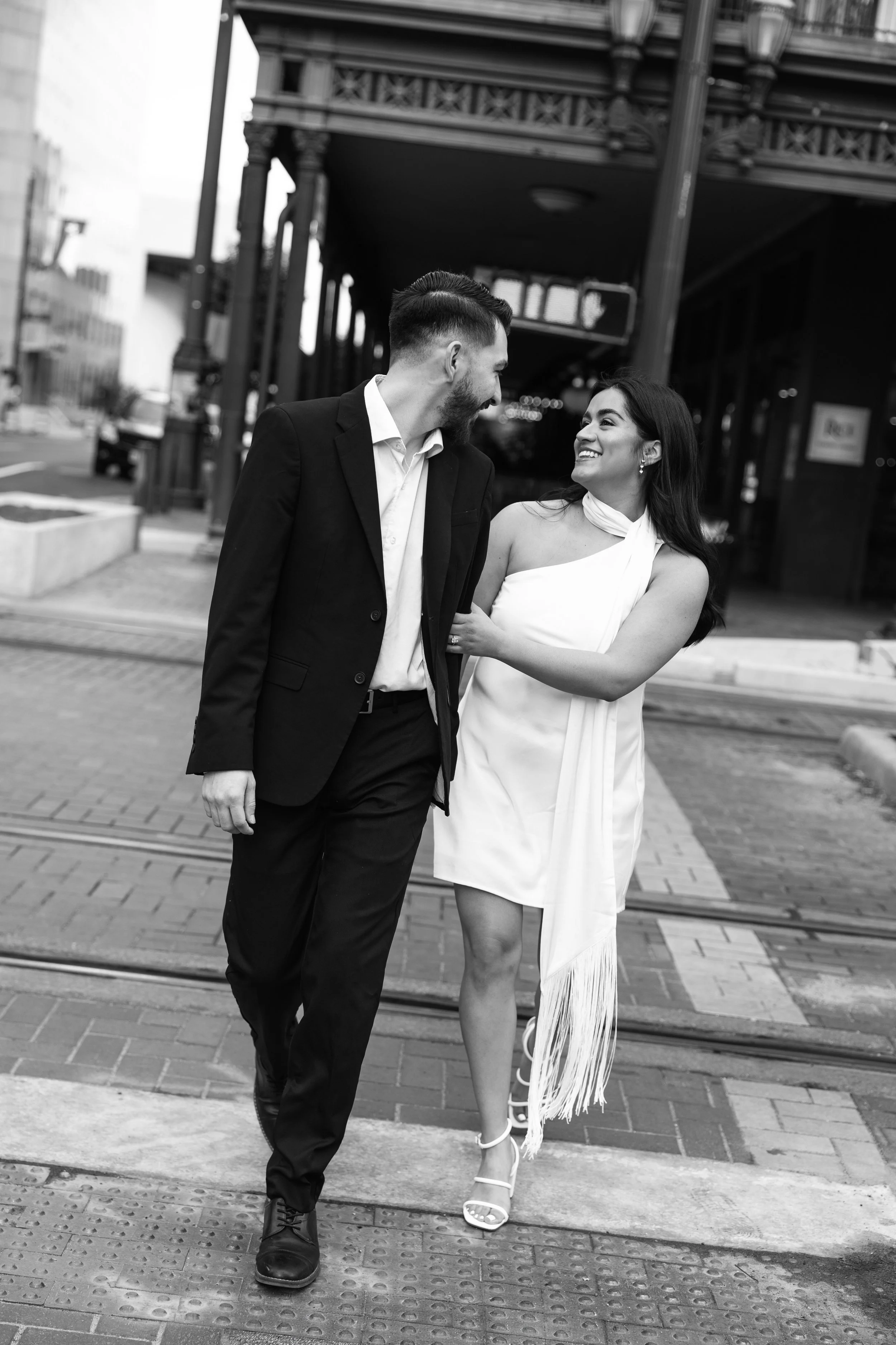 A couple walking together on a city street, smiling at each other. The man is wearing a suit and the woman is wearing a light-colored dress with high heels. They are under a structure with decorative columns and tram tracks are visible on the street.