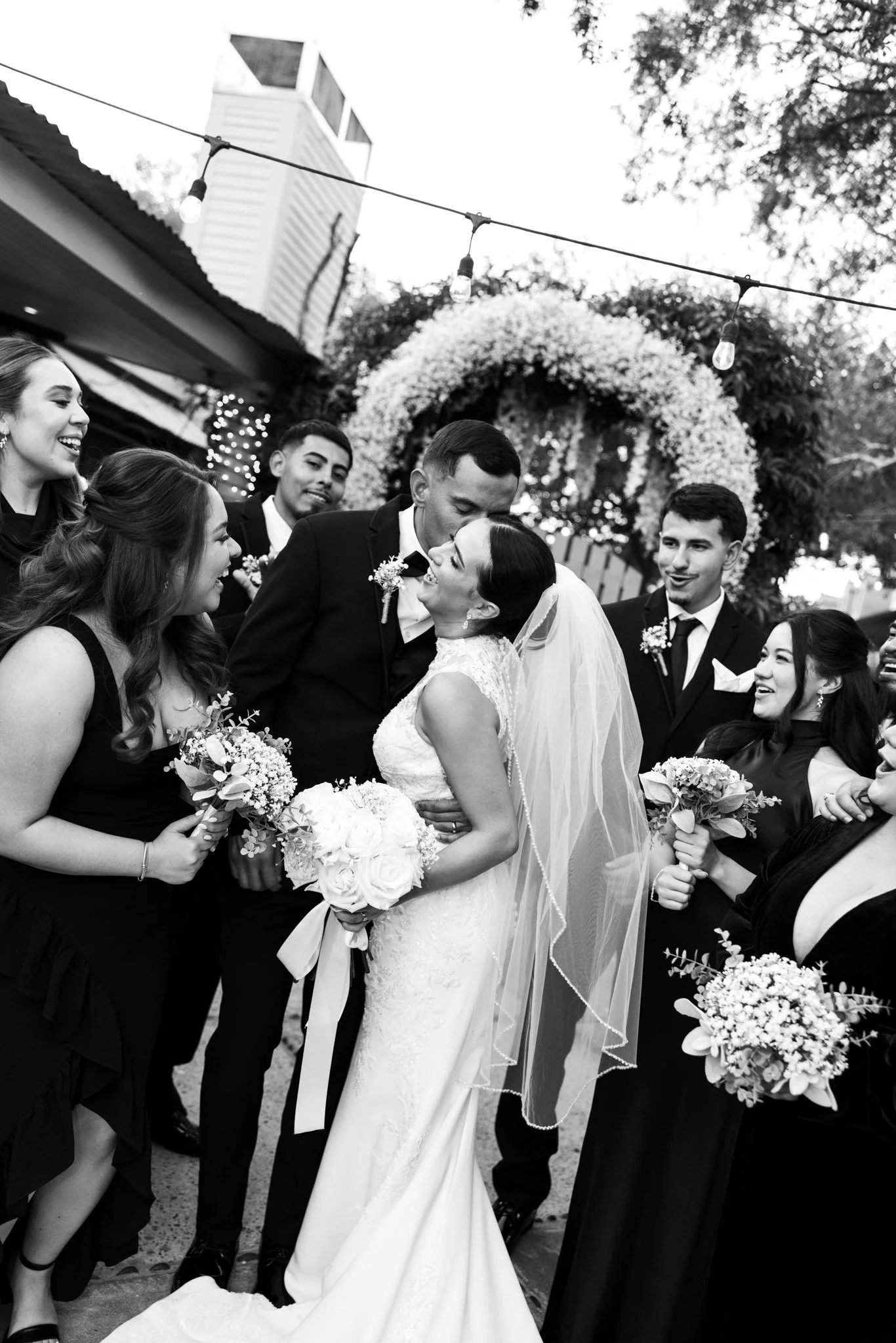 Black and white photo of a wedding scene with the bride and groom kissing, surrounded by bridesmaids and groomsmen holding bouquets, with floral decorations and string lights in the background.