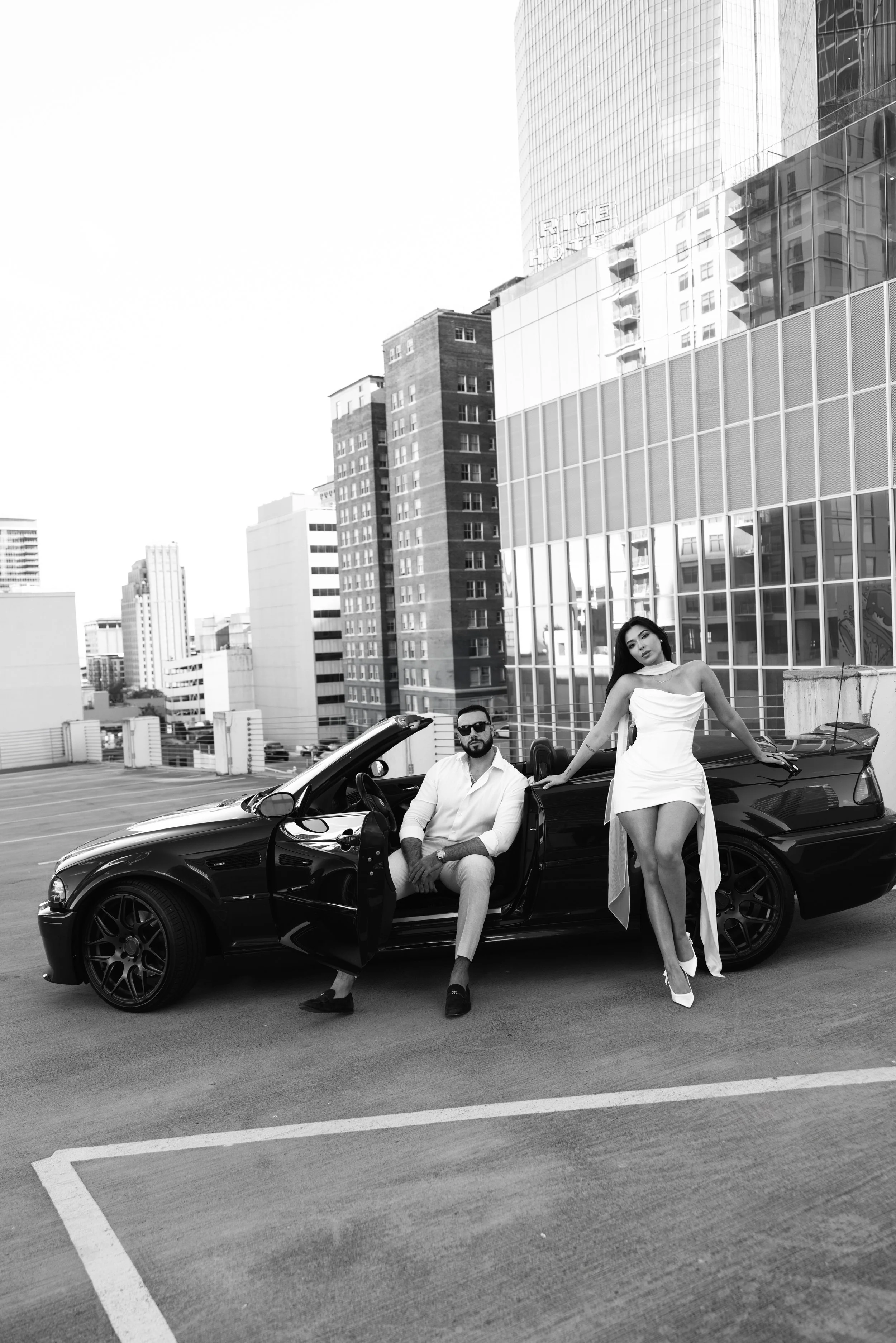 A man in a white shirt and light-colored pants sitting in a black convertible car with the door open, in an urban parking lot. A woman in an off-shoulder white dress and high heels stands beside him, leaning on the car with her right hand on the roof