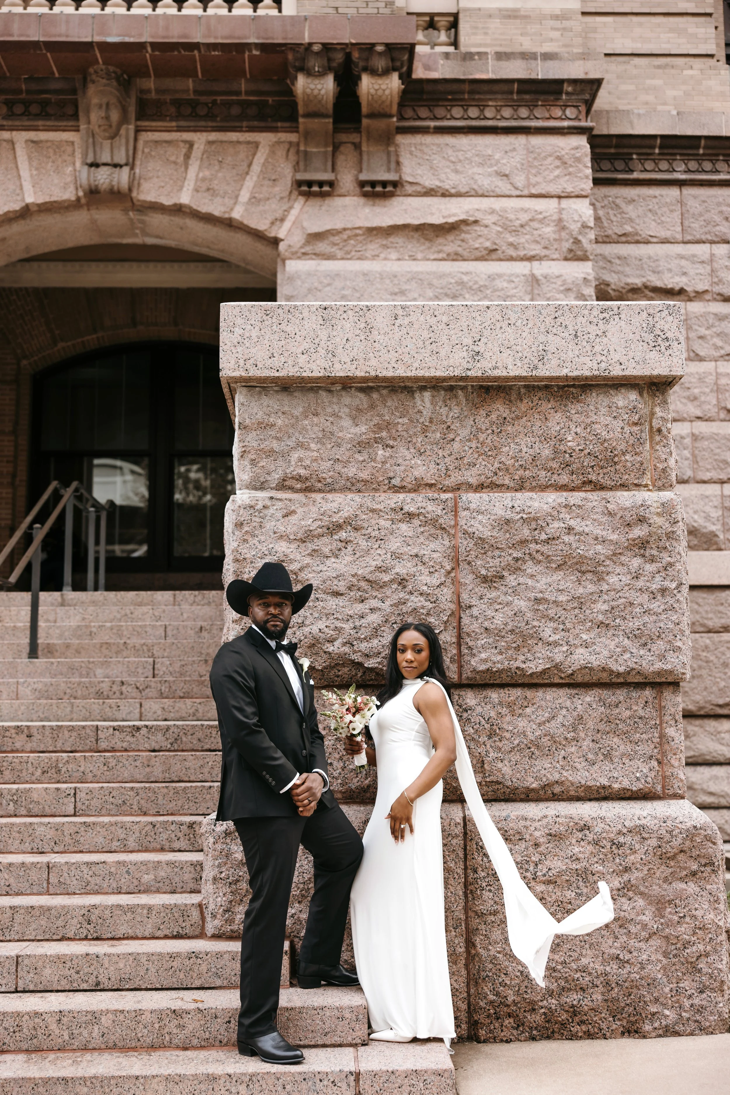 A bride and groom standing on steps in front of a stone building. The groom is wearing a black tuxedo with a cowboy hat, and the bride is in a white wedding gown holding a bouquet of flowers.
