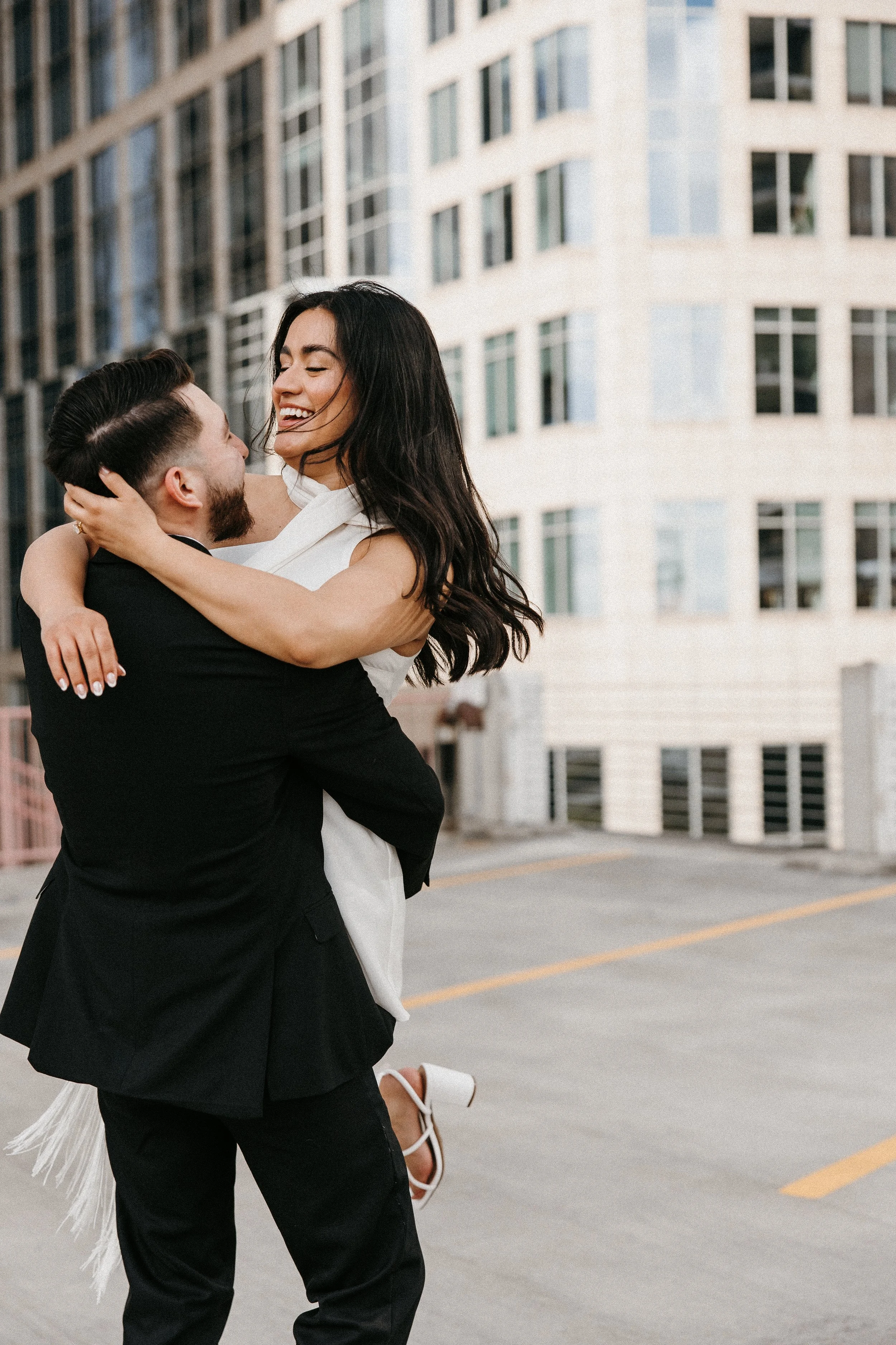 A man in a black suit and a woman in a white dress are embracing and smiling in an urban setting with high-rise buildings in the background.