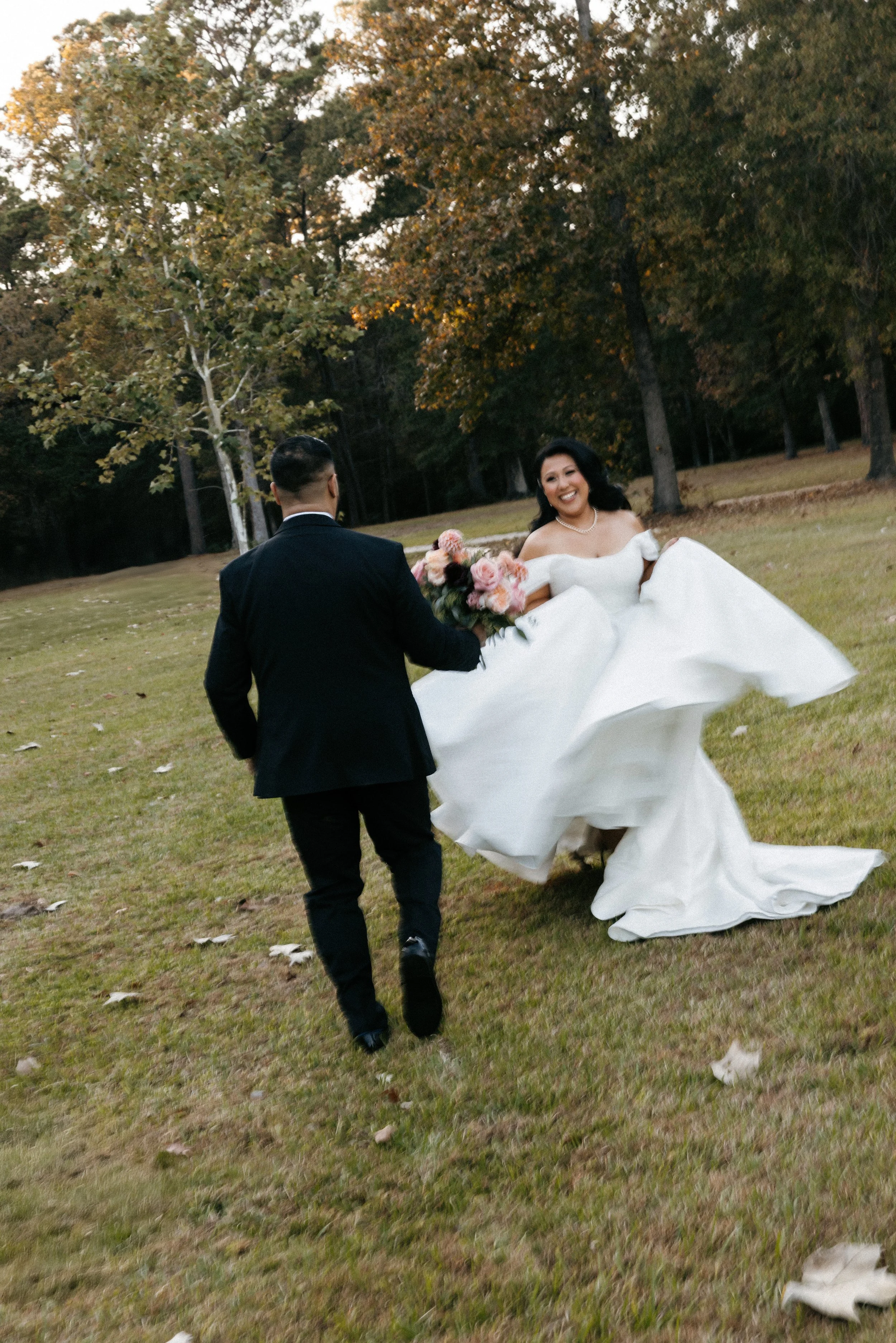 A bride in a white wedding dress and pearl necklace holding a bouquet of pink flowers, smiling as she walks in a grassy outdoor area with a groom in a black suit walking towards her, surrounded by trees with autumn leaves.