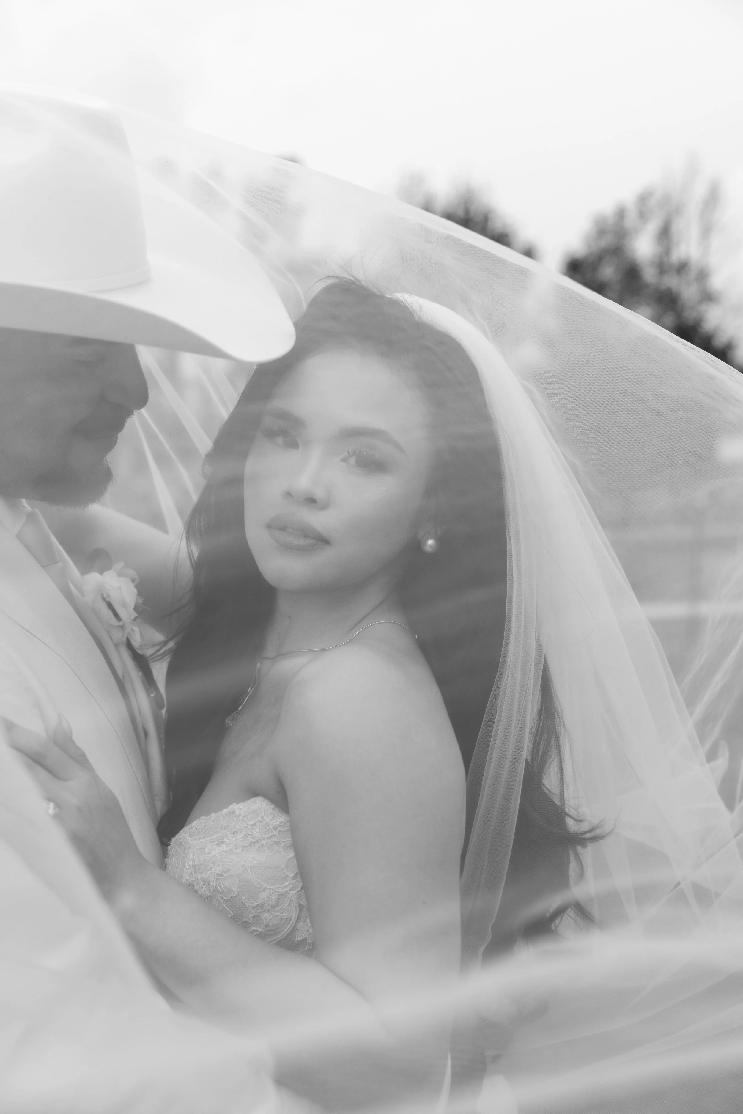 A black and white photo of a bride and groom close together, partially obscured by a veil, outside with trees in the background.