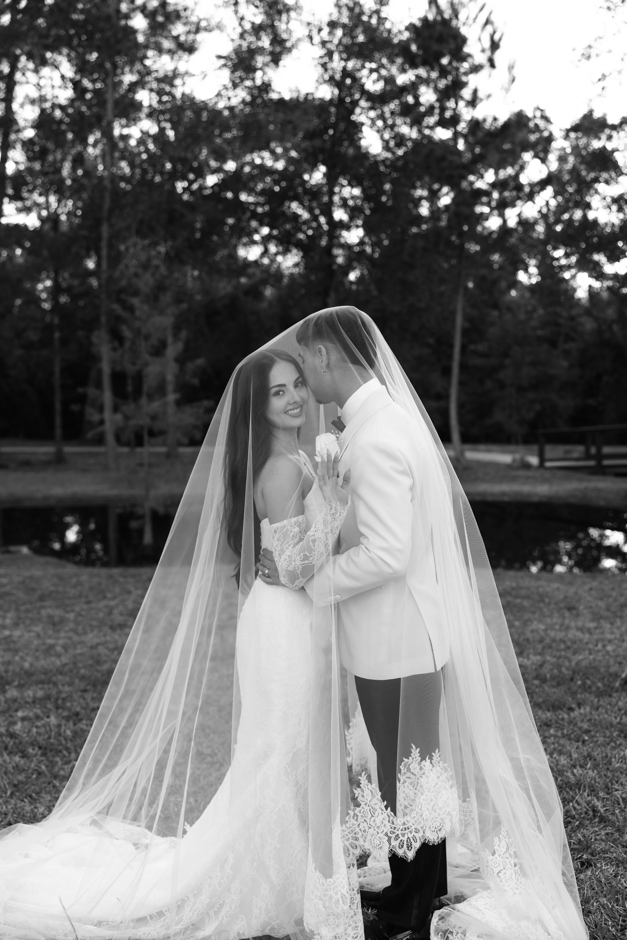 A bride and groom standing close together outdoors, with the bride smiling at the camera and the groom resting his head near her forehead, both covered by a sheer veil. The bride is holding a small flower, and trees are visible in the background.