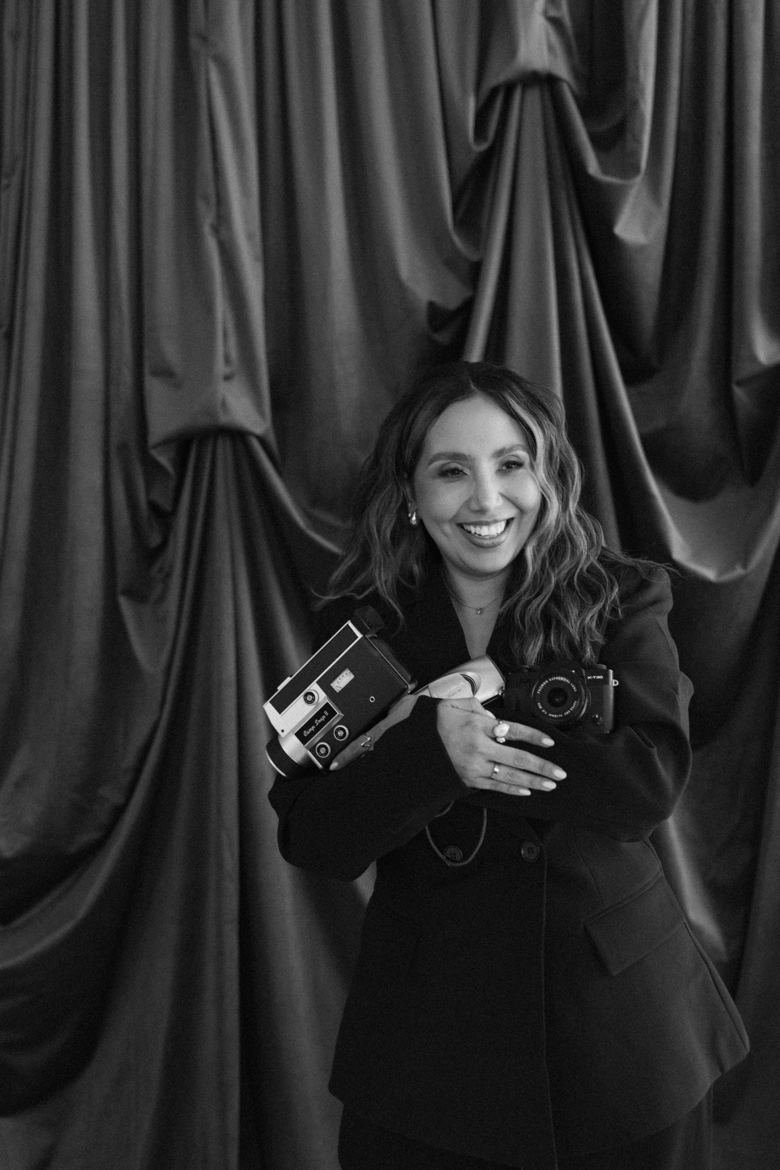 Black and white photo of a woman smiling, holding a camera, standing in front of a curtain.