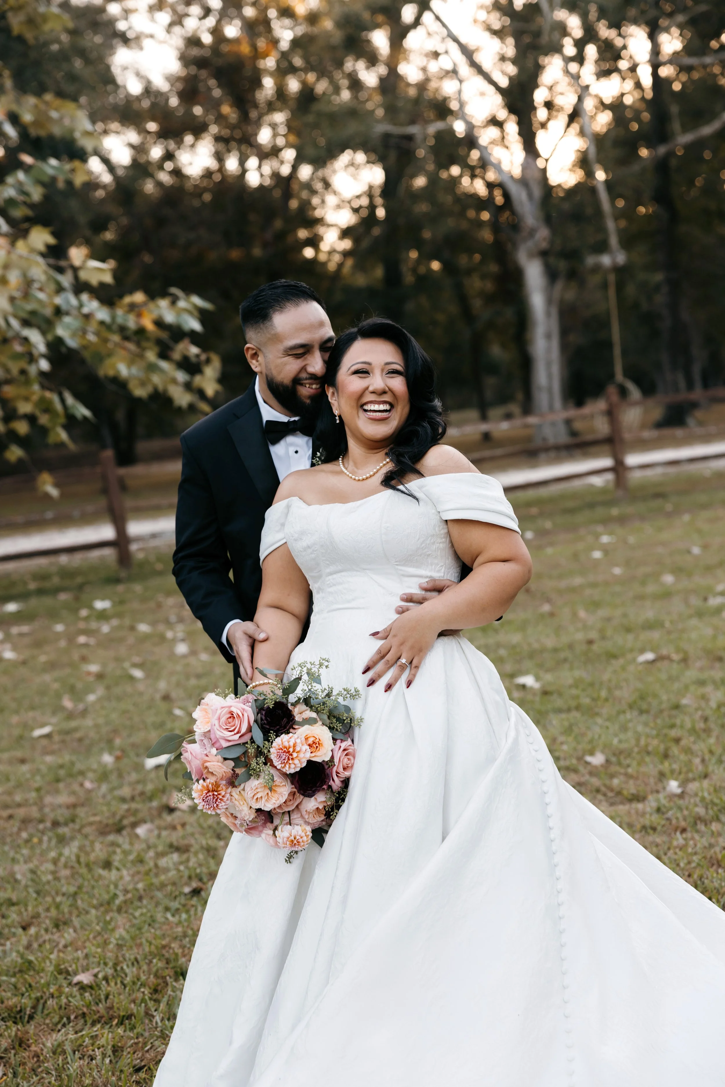 A happy bride and groom sharing a joyful moment outdoors during daylight, with trees and a wooden fence in the background. The bride is holding a bouquet of pink, peach, and dark purple flowers, wearing a white off-shoulder wedding gown and a pearl n