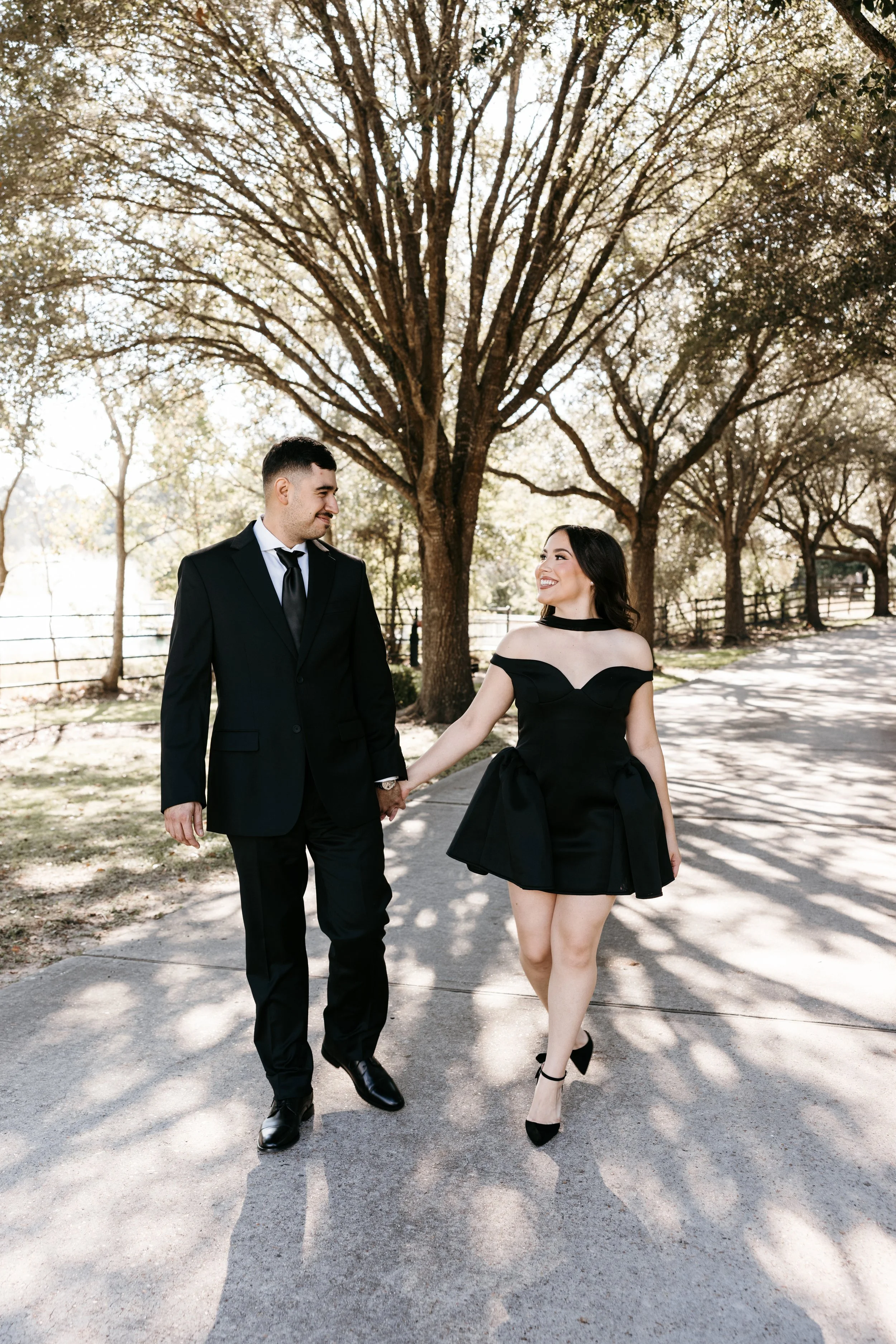 A young couple walking hand in hand outdoors on a sunny day, surrounded by trees and a park setting, dressed in formal attire, smiling and enjoying each other's company.