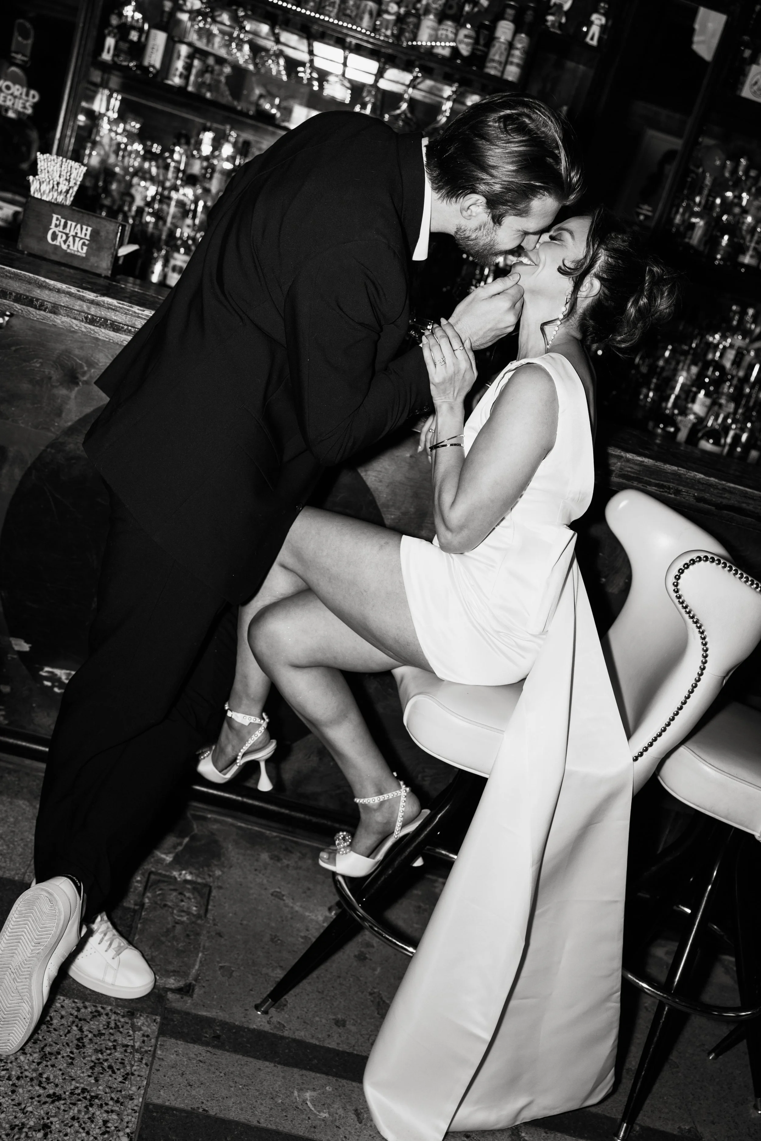 A couple sharing a romantic moment at a bar, with the man leaning over and holding the woman's face, both smiling, and the woman sitting on a barstool wearing high heels and a white dress.