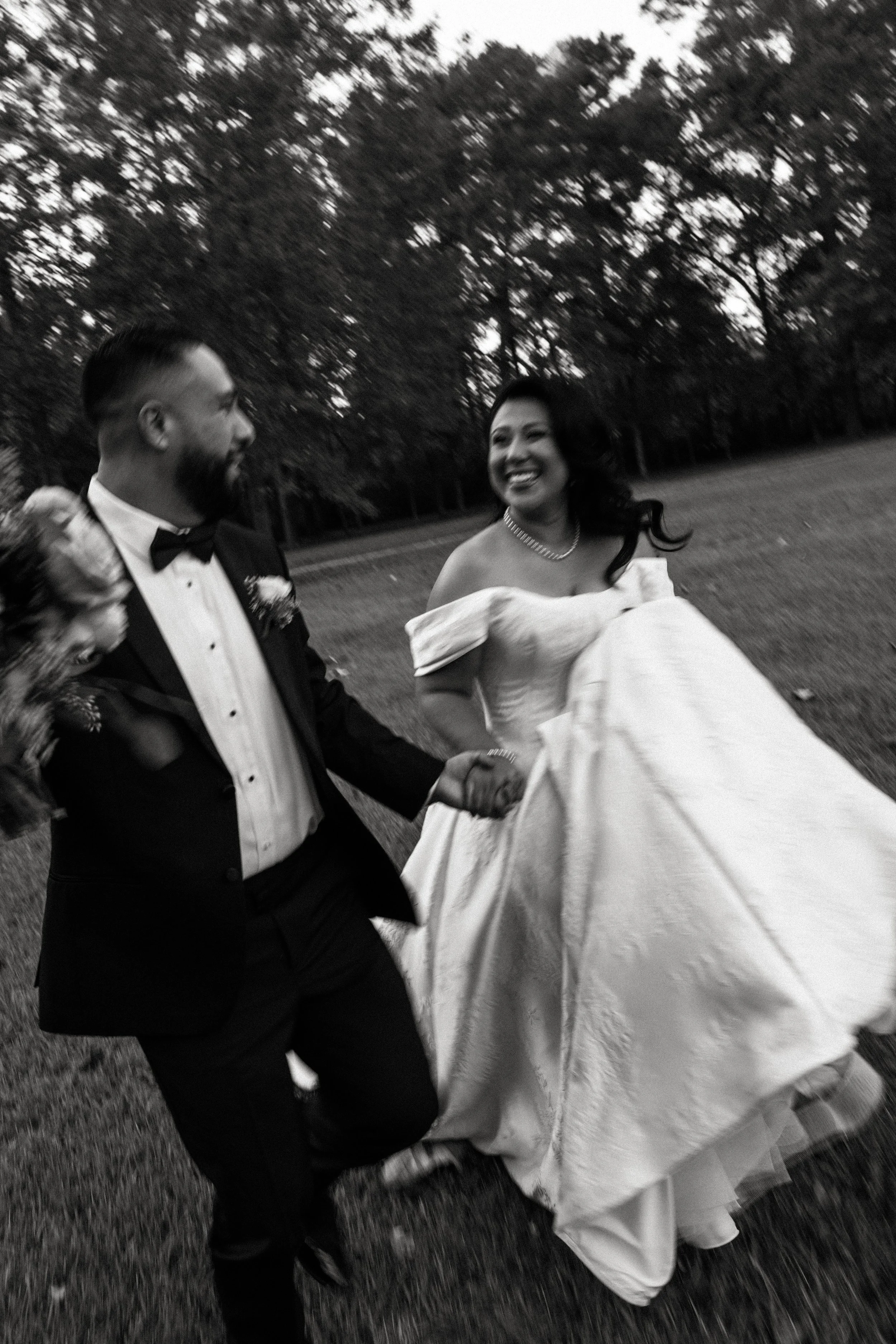 A bride and groom walking outdoors, holding hands, with trees in the background, the bride is smiling and wearing an off-shoulder wedding dress, the groom is wearing a tuxedo with a bow tie, and they are holding hands, possibly during their wedding.