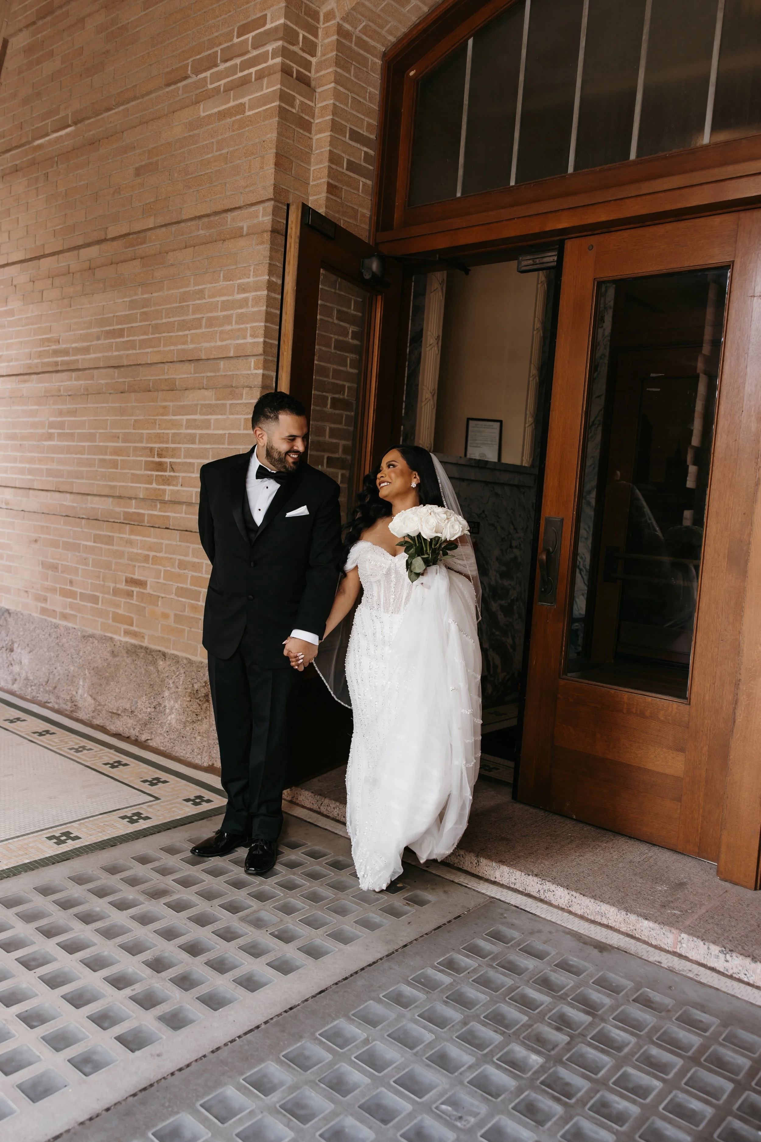 A newlywed couple stands outside a building entrance, holding hands and smiling at each other. The groom wears a black tuxedo with a bow tie and the bride wears a white wedding gown with a bouquet of white roses.