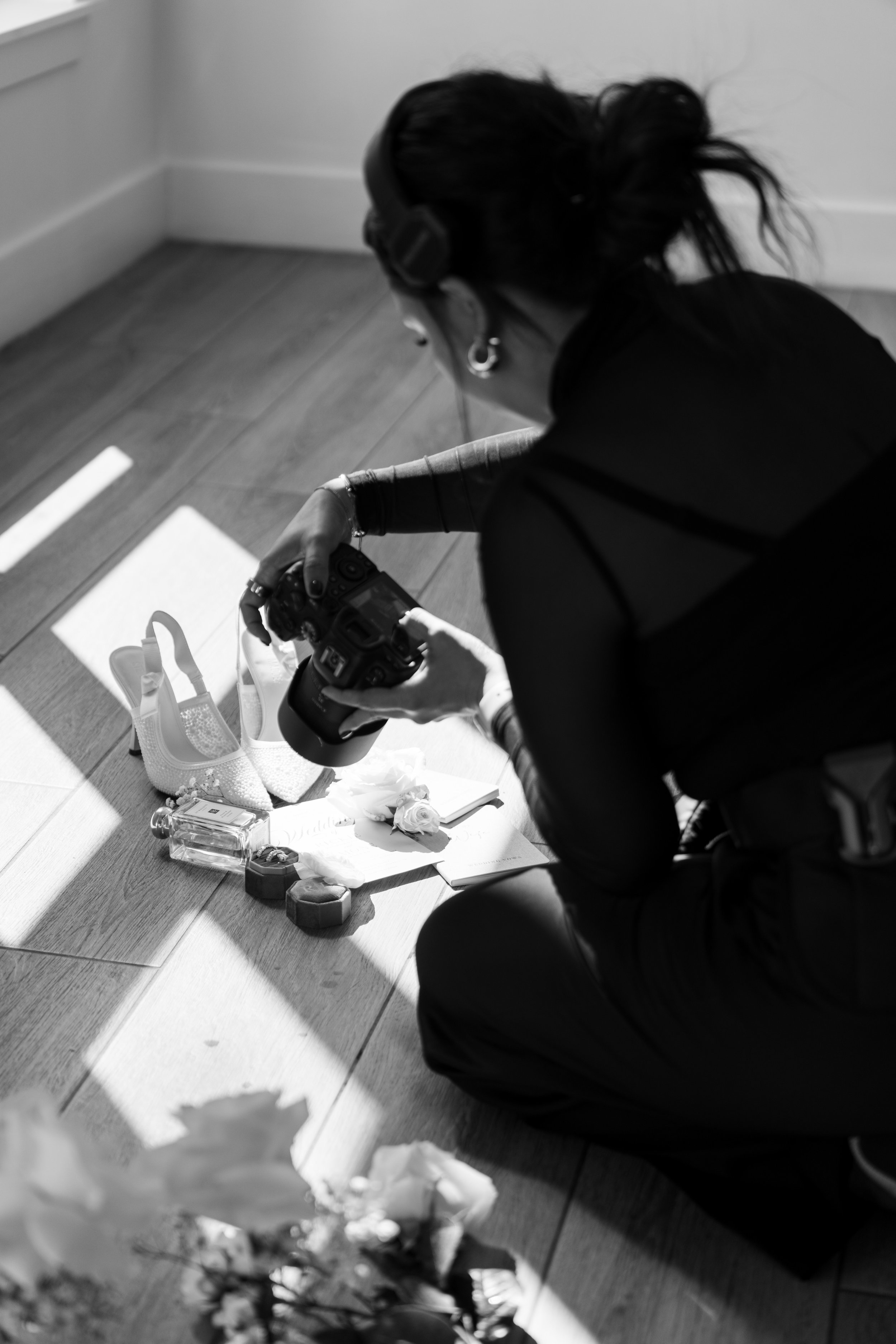 A person with dark curly hair, wearing headphones and earrings, is kneeling on a wooden floor, taking a photo of a white saddle with flowers, a small perfume bottle, and other objects laid out on a white sheet.
