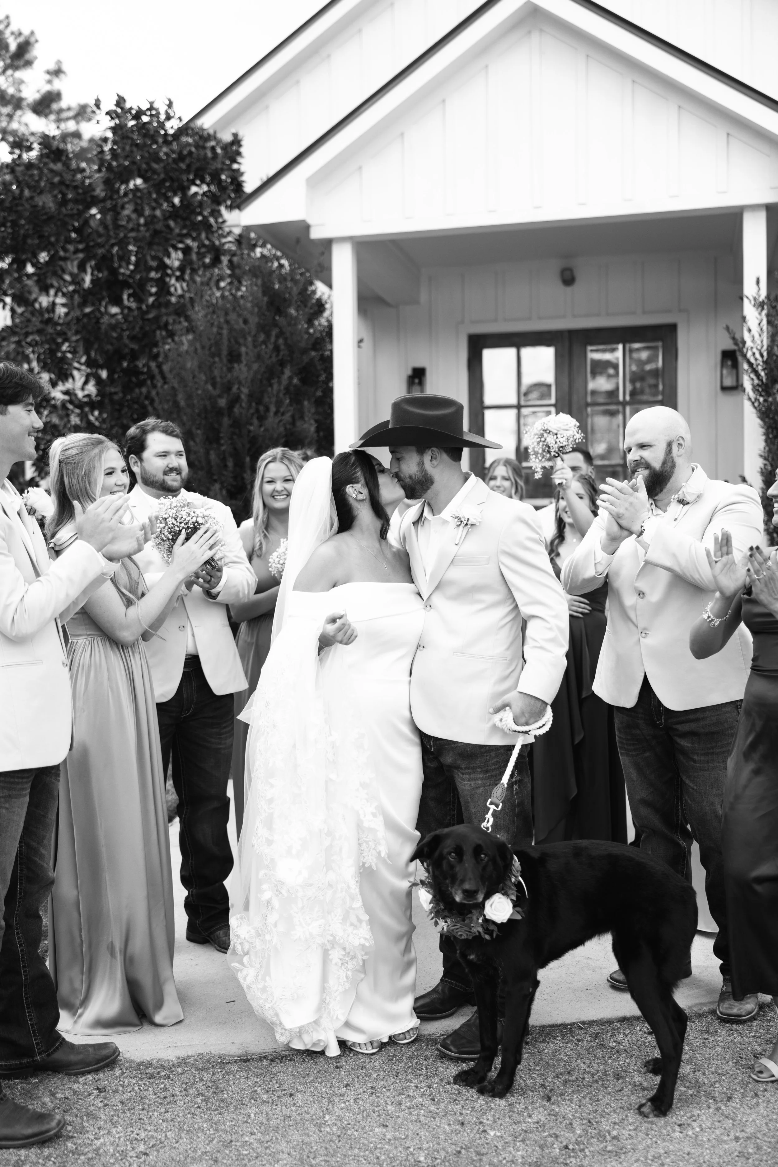 A black and white photo of a wedding ceremony outside with the bride and groom kissing. The bride is wearing a white dress and veil, and the groom is in a white suit with a wide-brimmed hat. They are surrounded by joyful guests, some holding bouquets