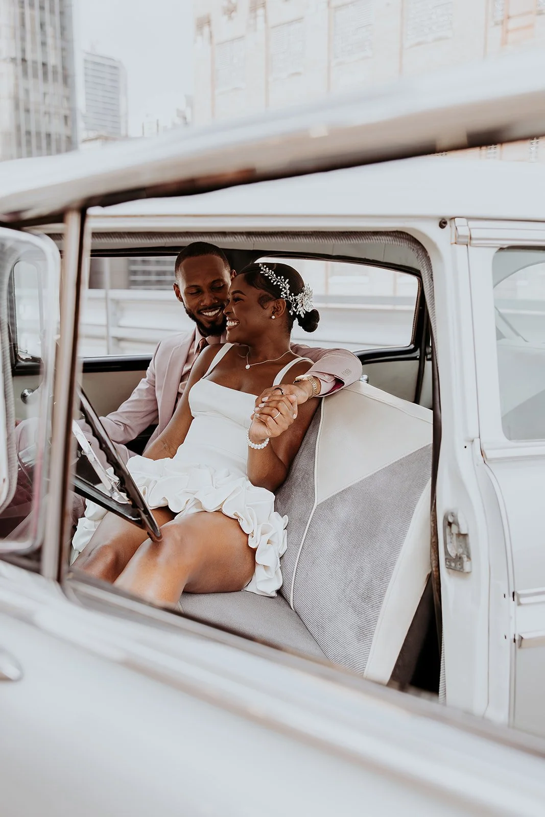 A newlywed couple happily sitting inside a vintage car, sharing a joyful moment and smiling at each other.