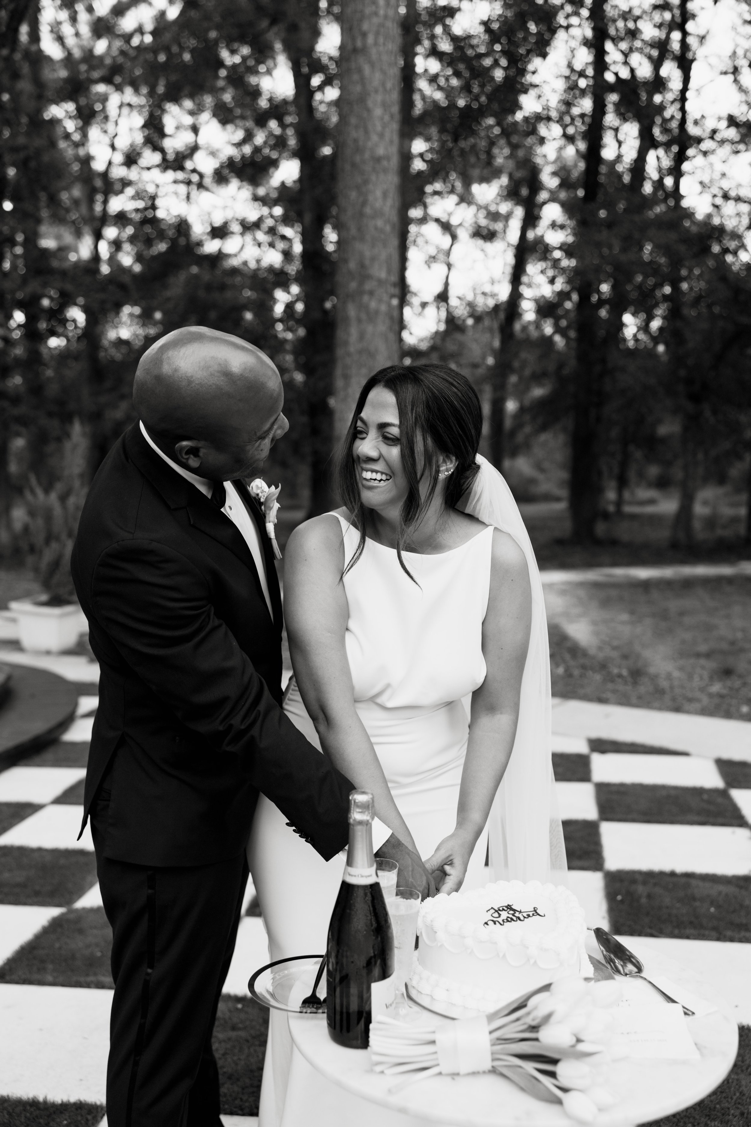 A black and white photo of a bride and groom smiling and holding hands during their wedding reception outdoors, with a checkered dance floor and wedding cake on a table nearby.