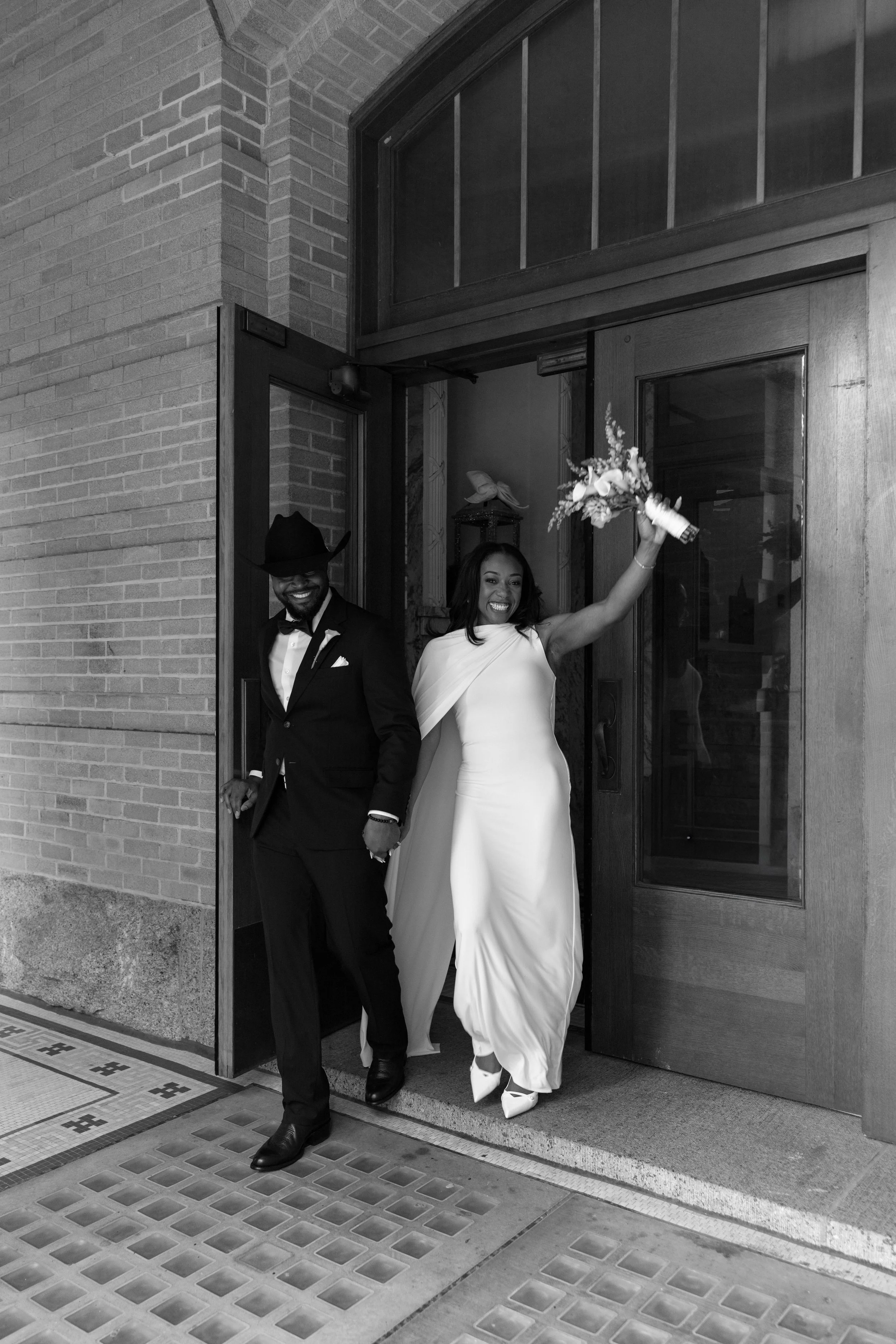 A joyful bride and groom newly married, exiting a building through an open wooden door, holding hands. The bride is wearing a white wedding gown, and the groom is dressed in a tuxedo with a cowboy hat. She is holding a bouquet of flowers.