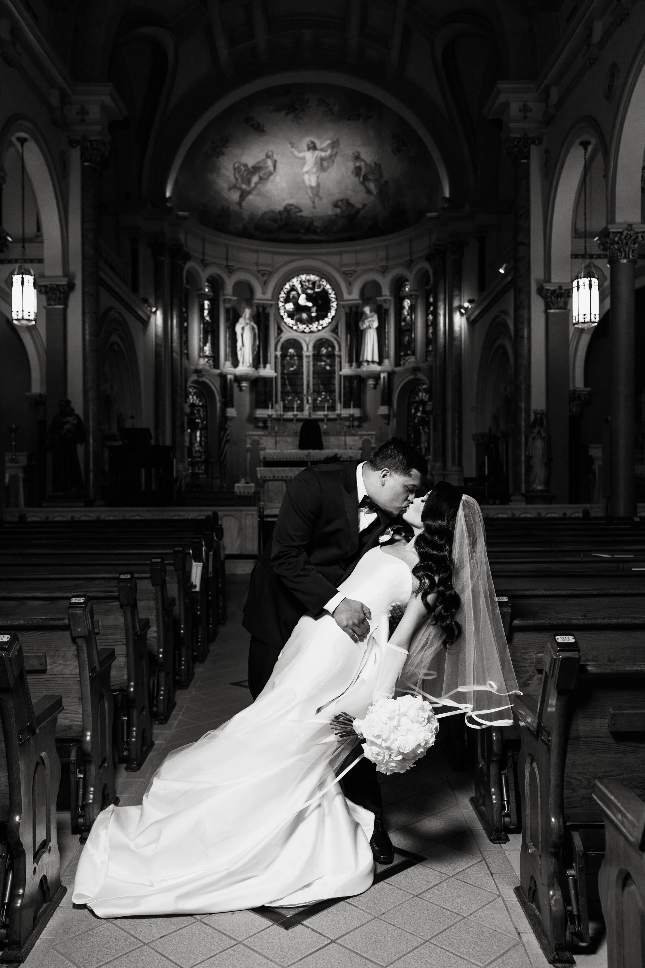 A bride and groom kissing inside a church with an ornate altar, religious statues, and a large painting of Jesus Christ on the ceiling, black and white photo.