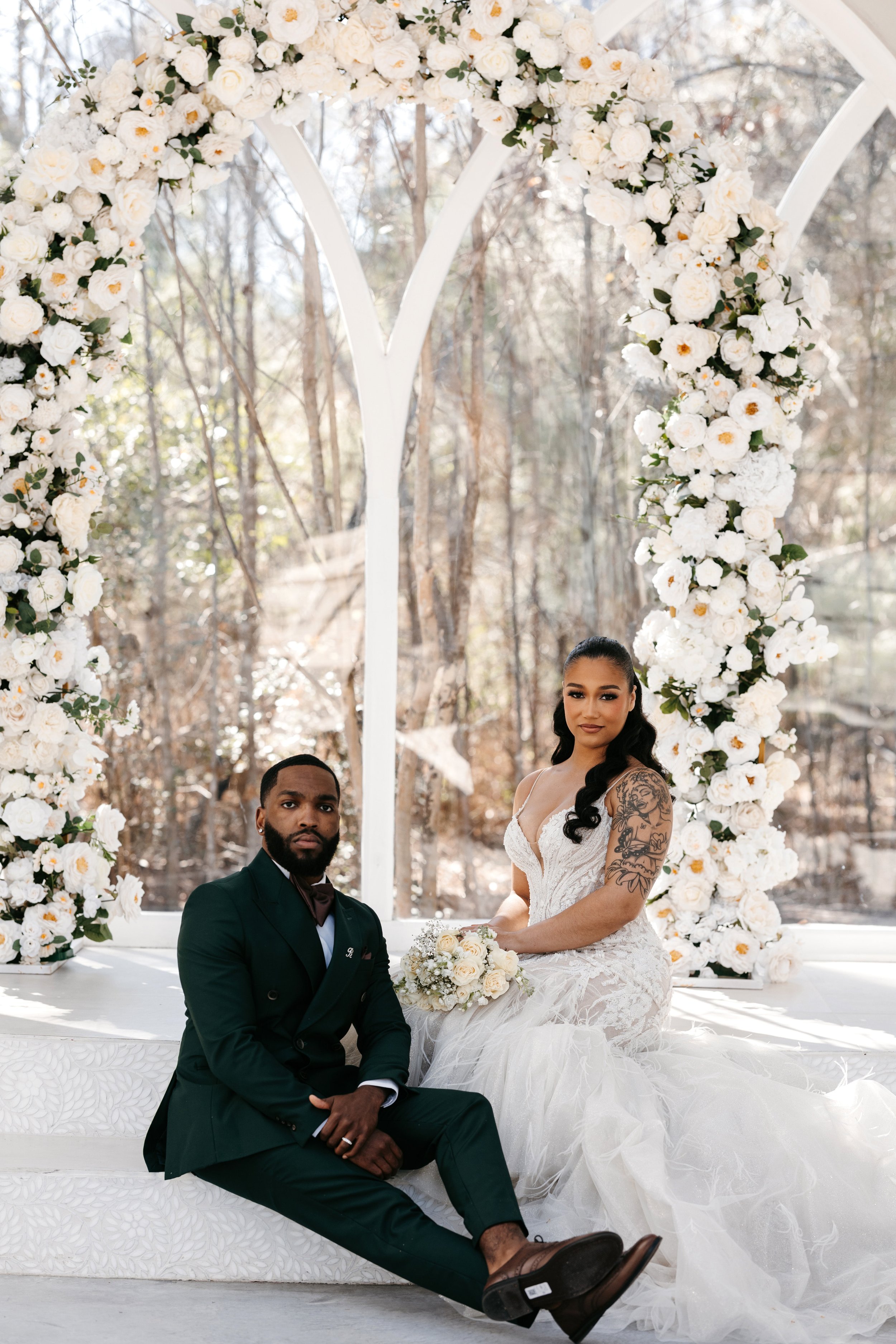 A wedding couple sitting on a white decorated platform under a floral arch, with a wooded outdoor background. The bride has dark hair, tattoos on her arm, and wears a white wedding dress. The groom has a beard, short hair, and wears a dark green suit