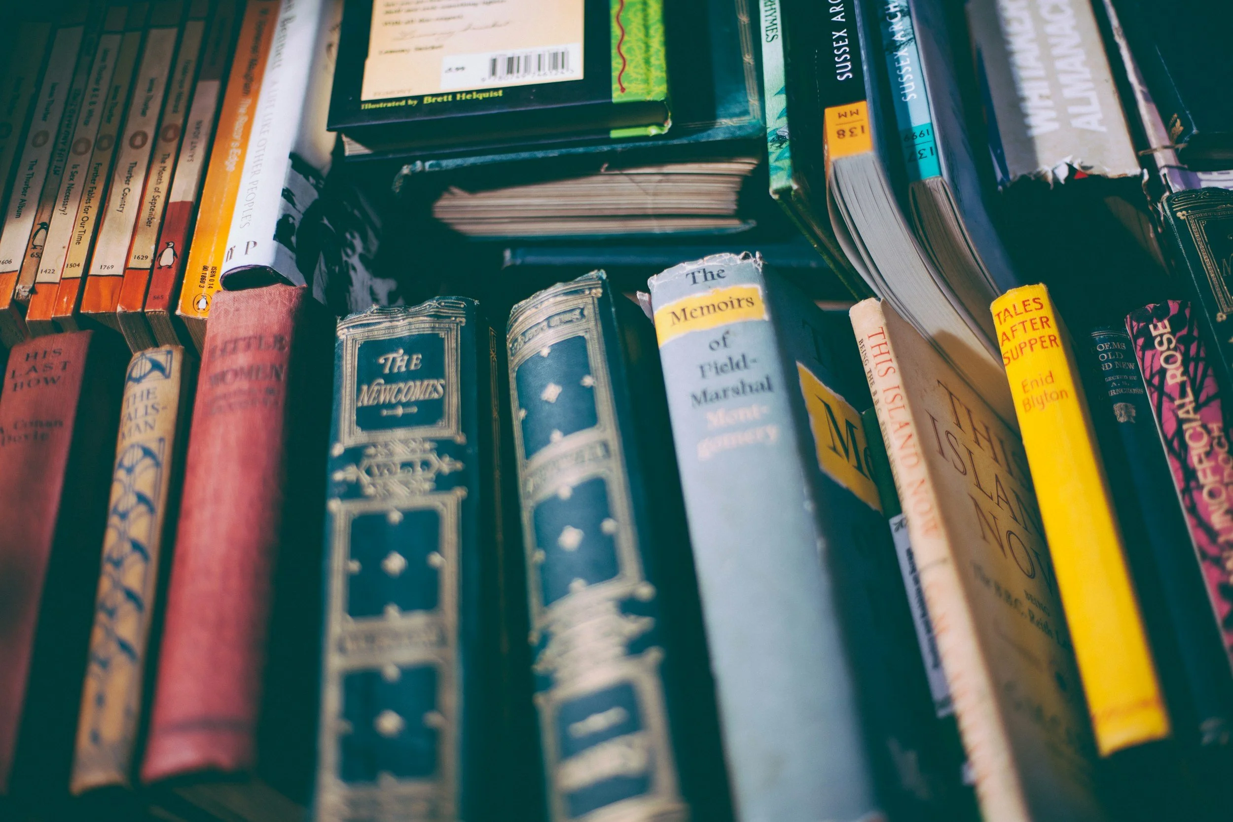 Close-up of a bookshelf with various books, including titles like 'The Newcomes,' 'Memoirs of Field Marshal Montgomery,' and 'Tales After Supper,' with some books stacked horizontally on top of others.