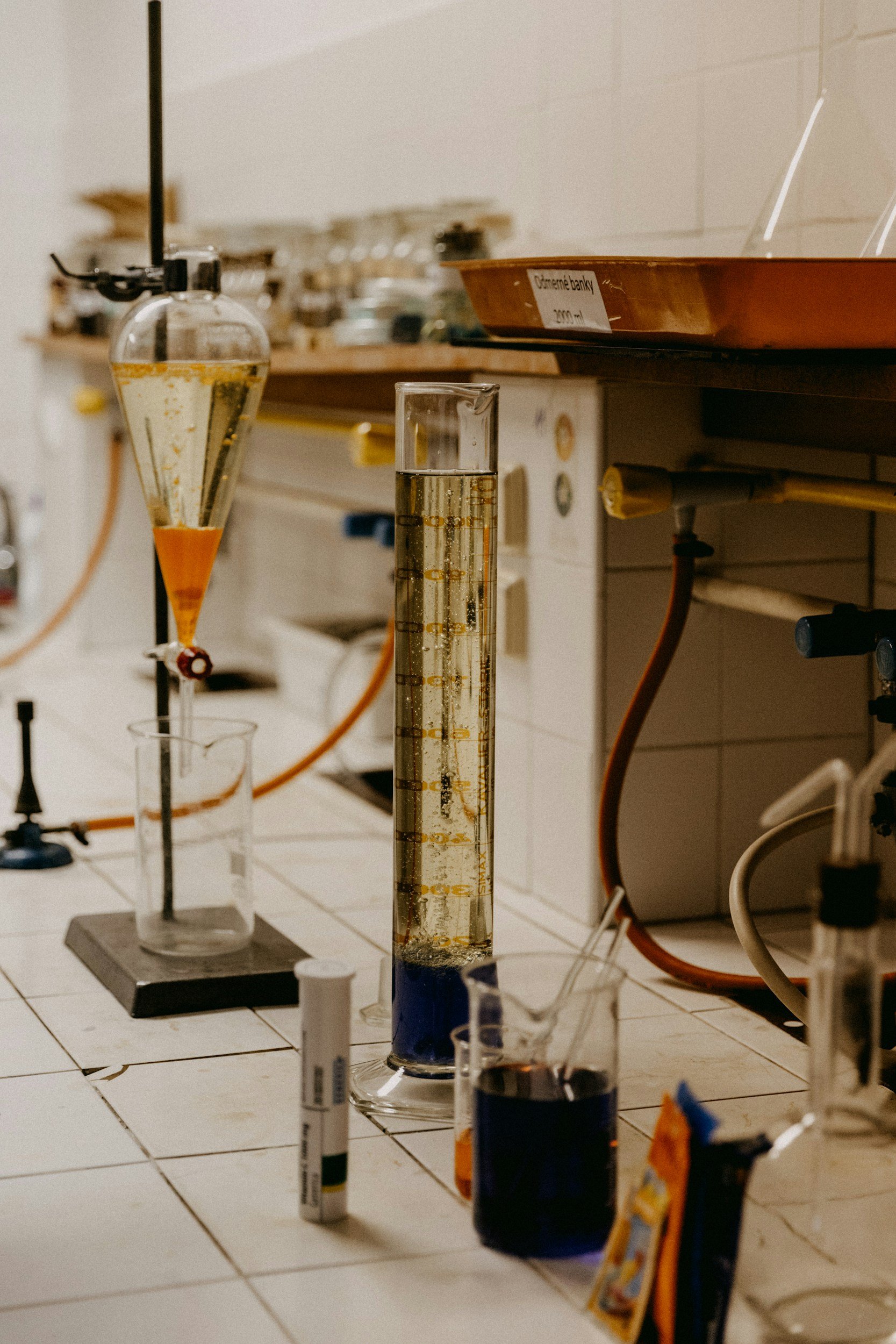 Laboratory setup with glassware containing colorful liquids, including graduated cylinders, beakers, and a funnel, on a tiled surface.