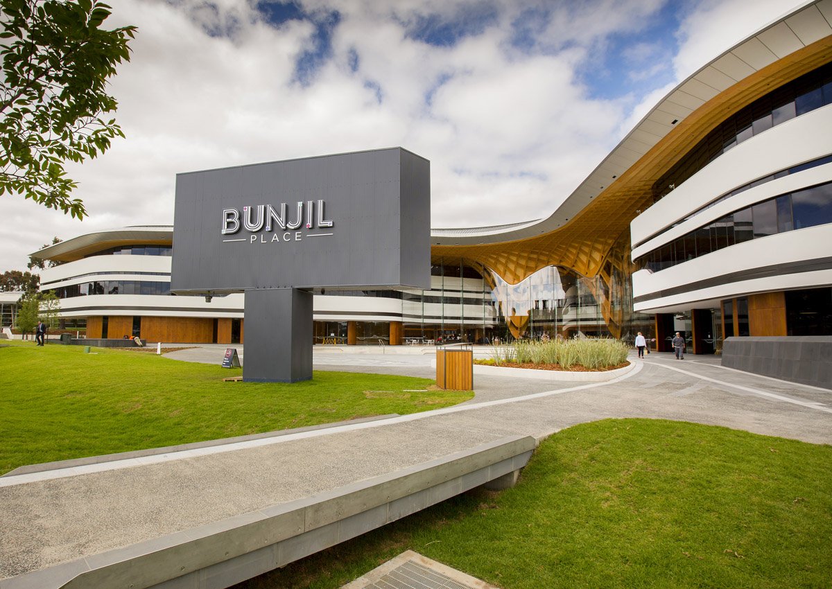 External view of Bunjil Place, a modern building with curved architecture, glass windows, and wooden accents, under a partly cloudy sky.