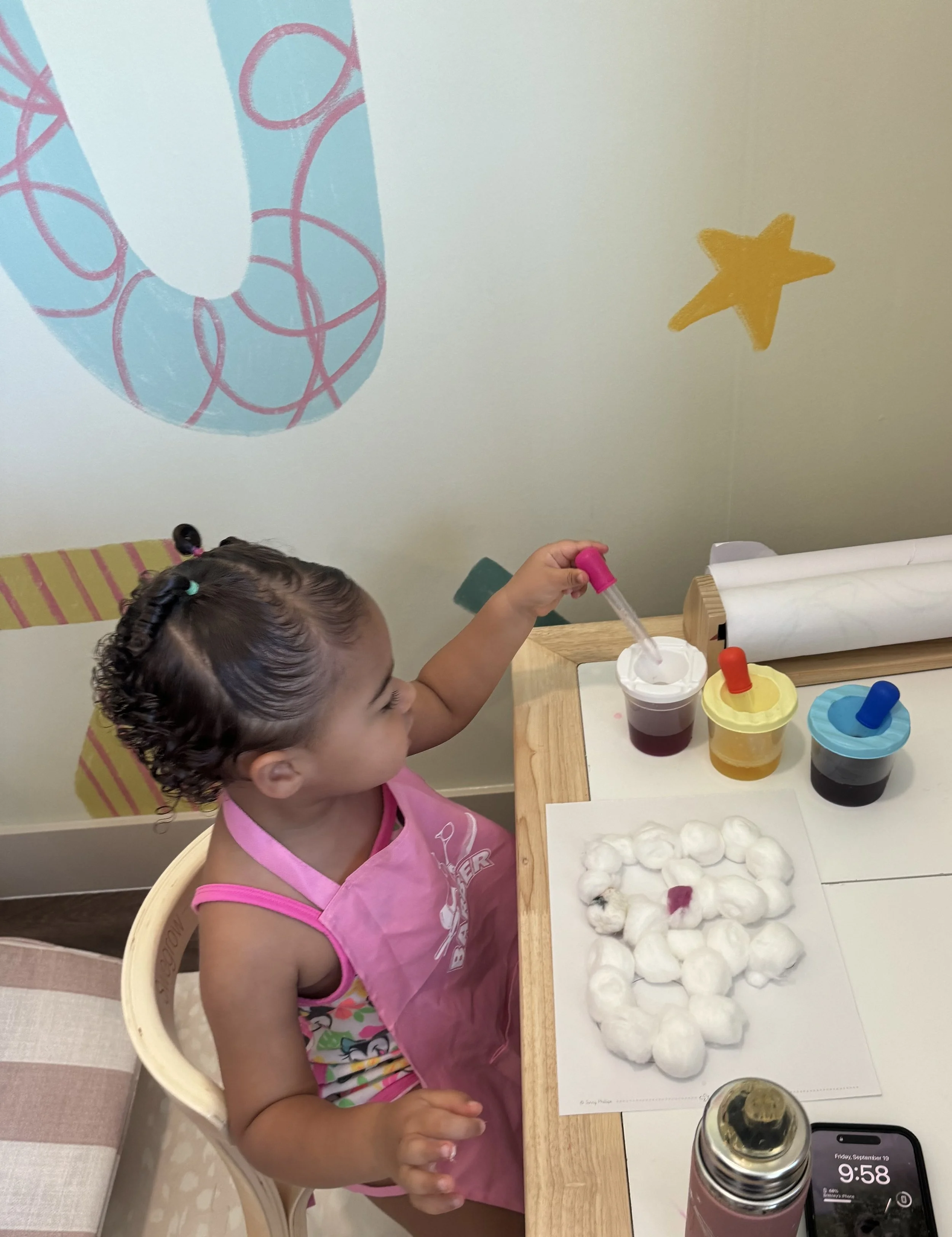 A young girl wearing a pink apron is sitting at a table, engaging in a cotton candy art activity with colored dye. She is holding a stick with a dropper, adding dye to the cotton candy, which is arranged on a paper sheet. On the table are three containers with differently colored dyes, a roll of paper towels, a pink water bottle, and a smartphone displaying the time 9:58. The background wall has colorful decorations, including a large blue and pink element and a yellow star.
