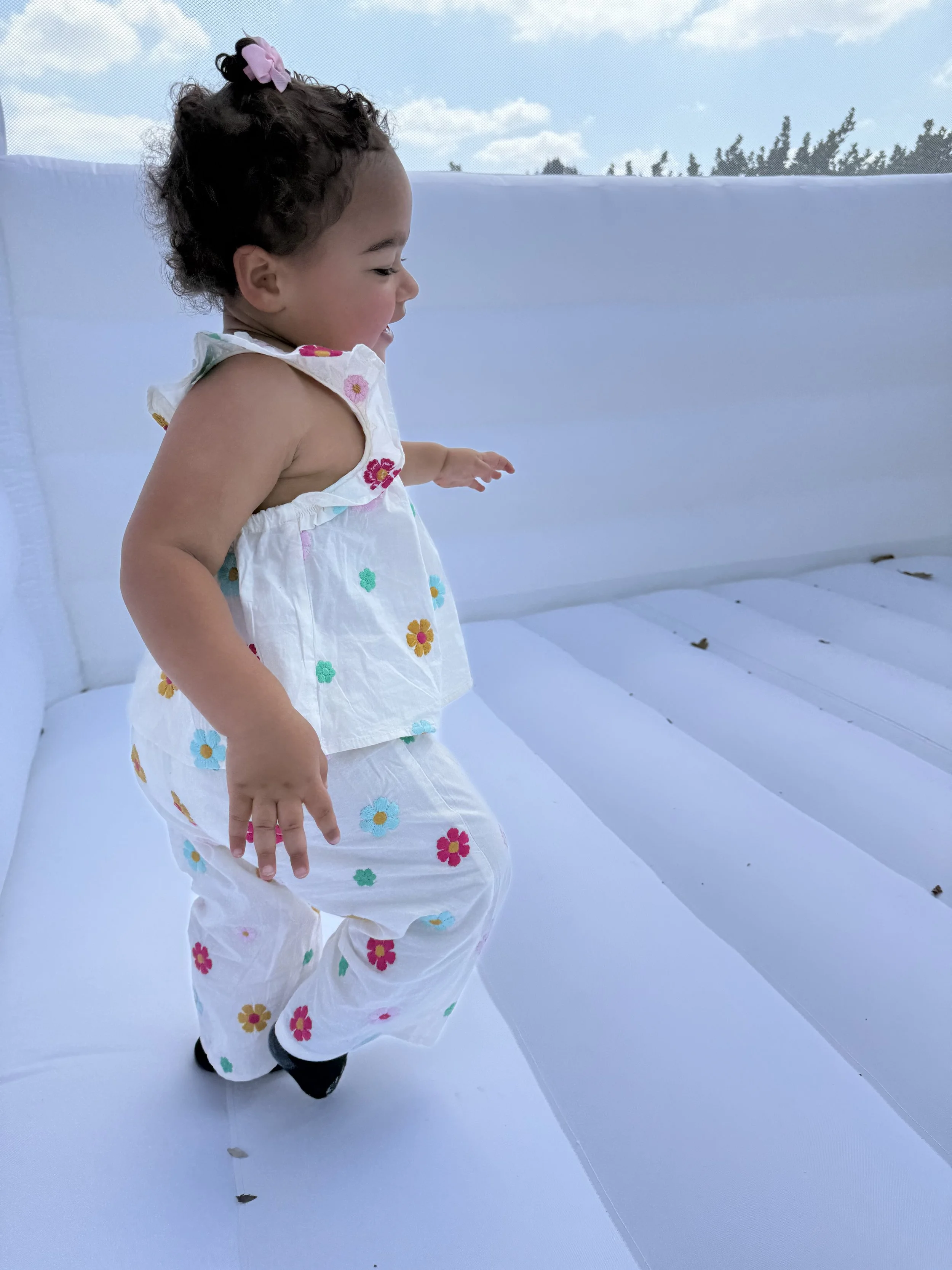 Young girl with dark curly hair, wearing a white floral outfit, jumping on an outdoor inflatable mattress or bounce house, with blue sky and trees in the background.