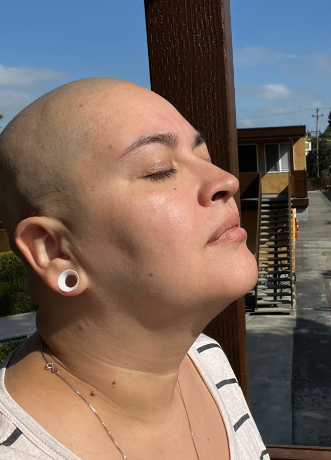 Close-up of a woman with a shaved head and ear gauges, relaxing with eyes closed outdoors on a sunny day