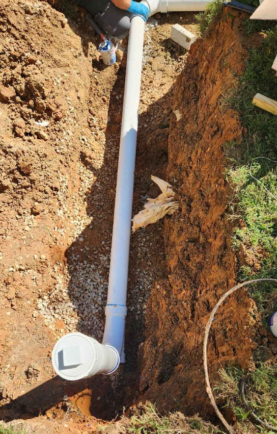 An underground plumbing pipe installation in a soil trench, with a white PVC pipe connected to a smaller black pipe and a white vent cap at the end. sewage drain, septic repair, plumber
