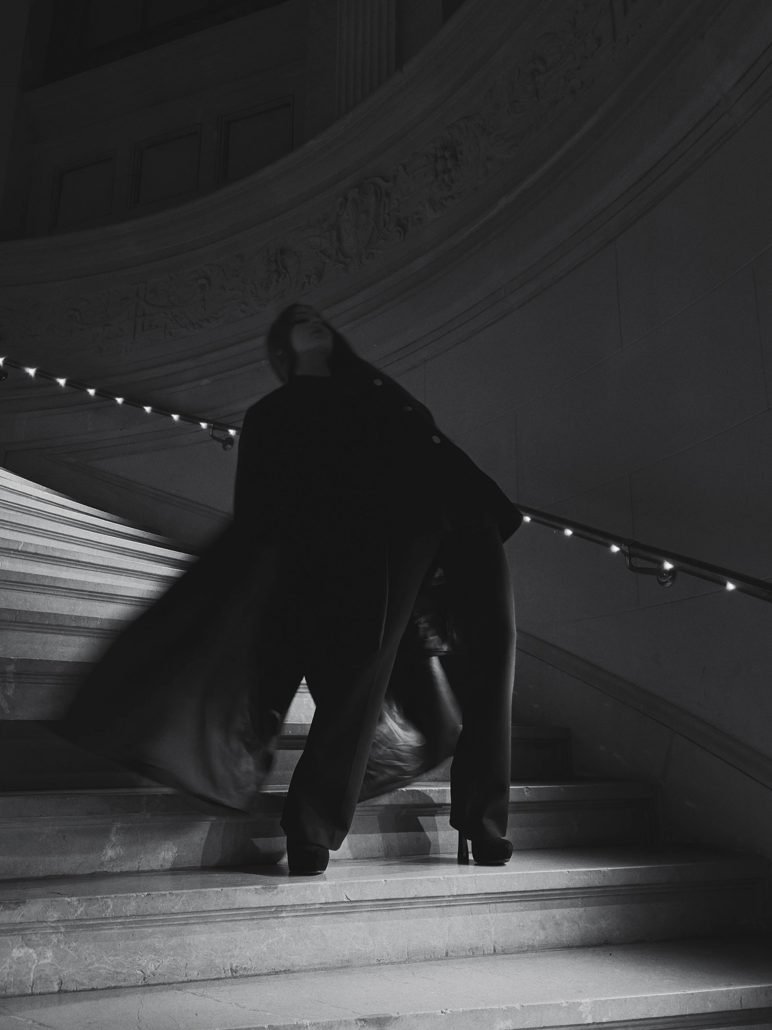 Une personne en talons hauts descendre un escalier en marbre dans un bâtiment élégant, éclairé faiblement, avec une rampe lumineuse et des détails décoratifs ornés au plafond.