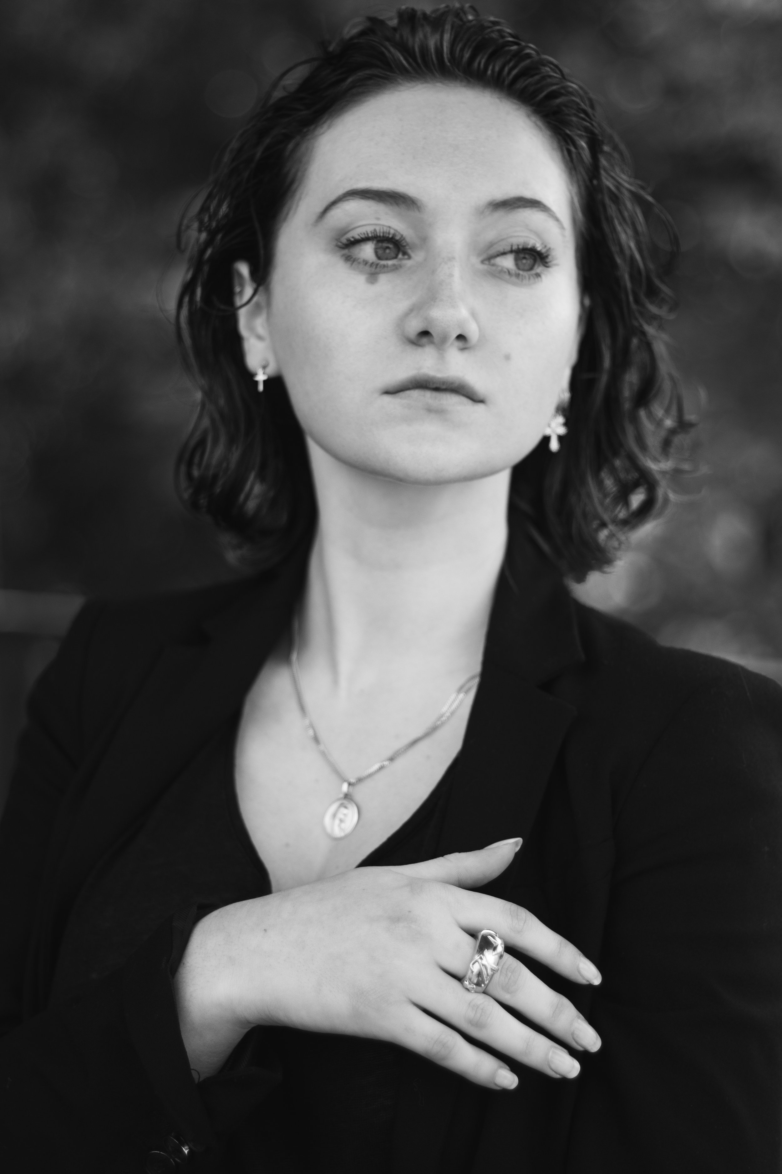 Portrait en noir et blanc d'une femme avec des cheveux ondulés, portant des boucles d'oreilles en forme de croix, un collier avec un médaillon et une bague, regardant vers la droite, fond flou.