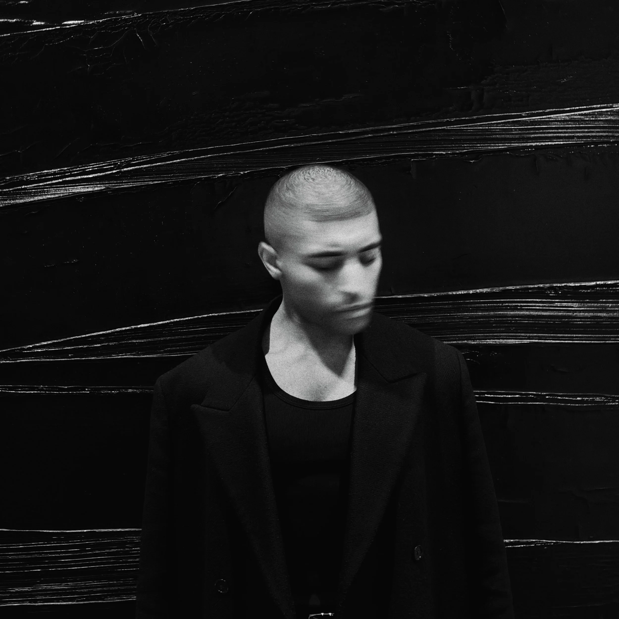 Un jeune homme avec une tête rasée, en costume noir, regardant vers le bas, devant un mur noir avec des textures et lignes blanches.
