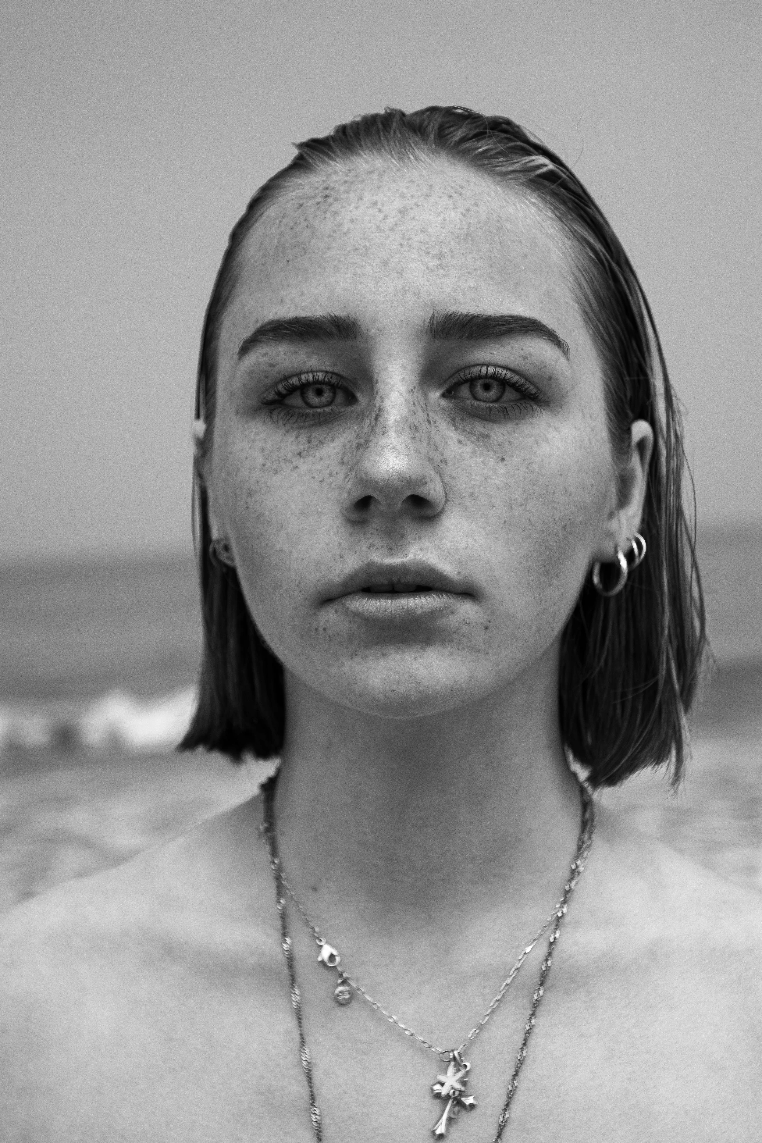 Un portrait en noir et blanc d'une jeune femme avec des cheveux courts, portant des boucles d'oreilles et plusieurs colliers, devant un fond de plage.