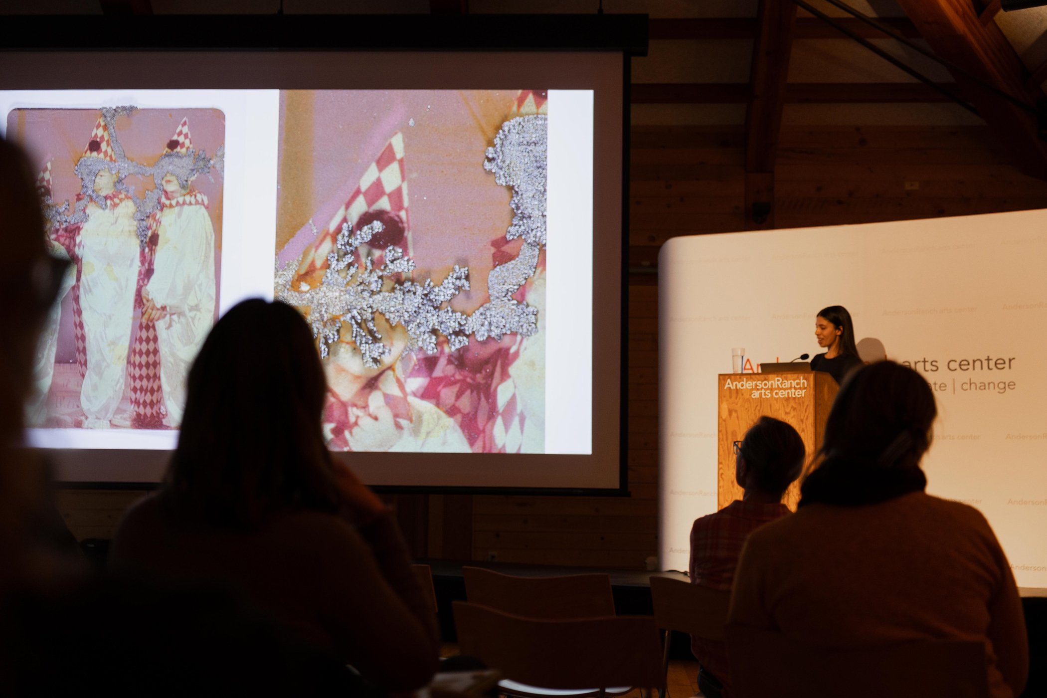 Karinne Smith presents at a podium at the Anderson Ranch Arts Center. A large screen displays an artwork featuring three clowns in costumes with checkered patterns.