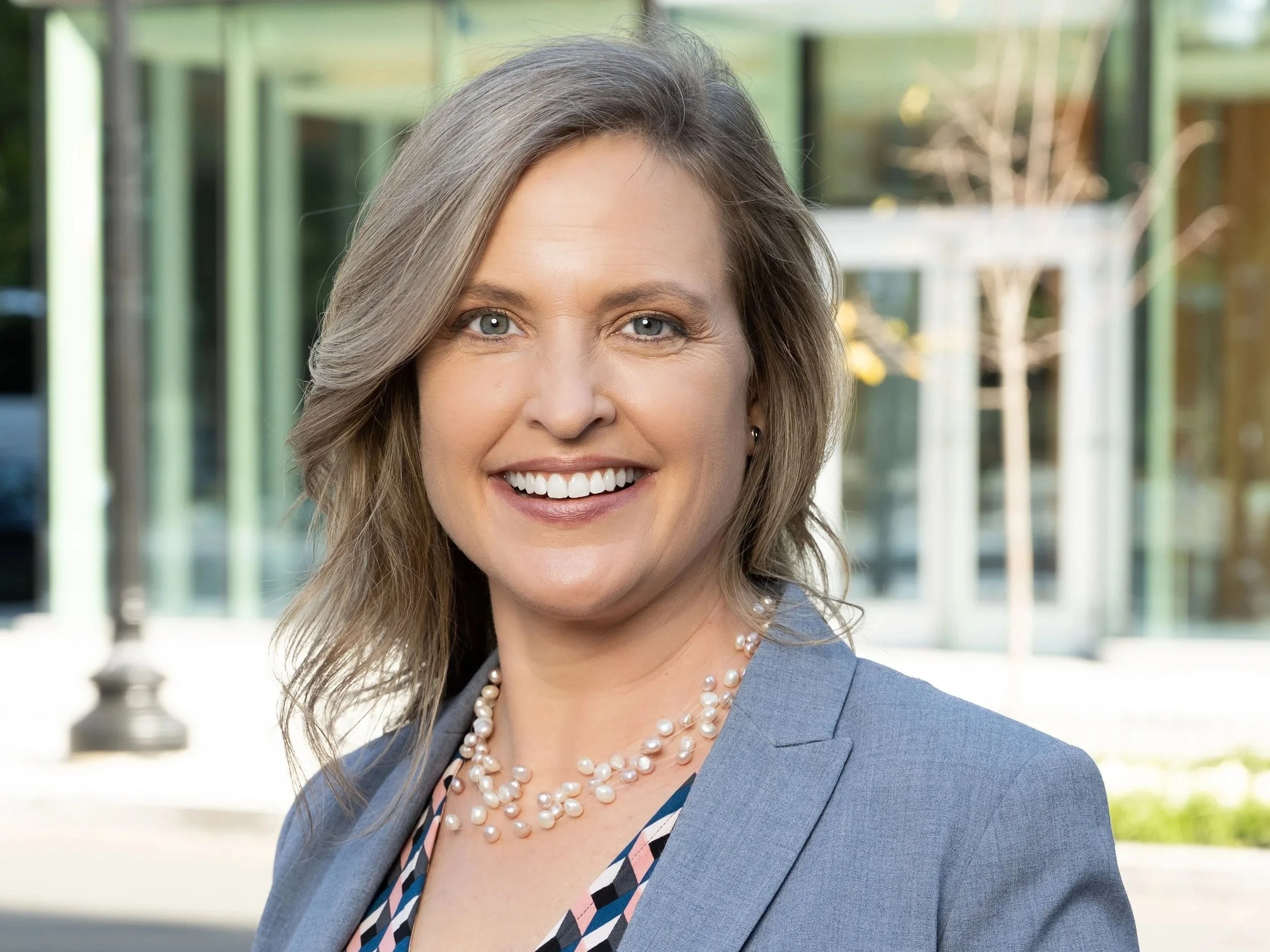 Portrait of a smiling woman in business attire with a blazer and pearl necklace outside in front of a modern glass building.