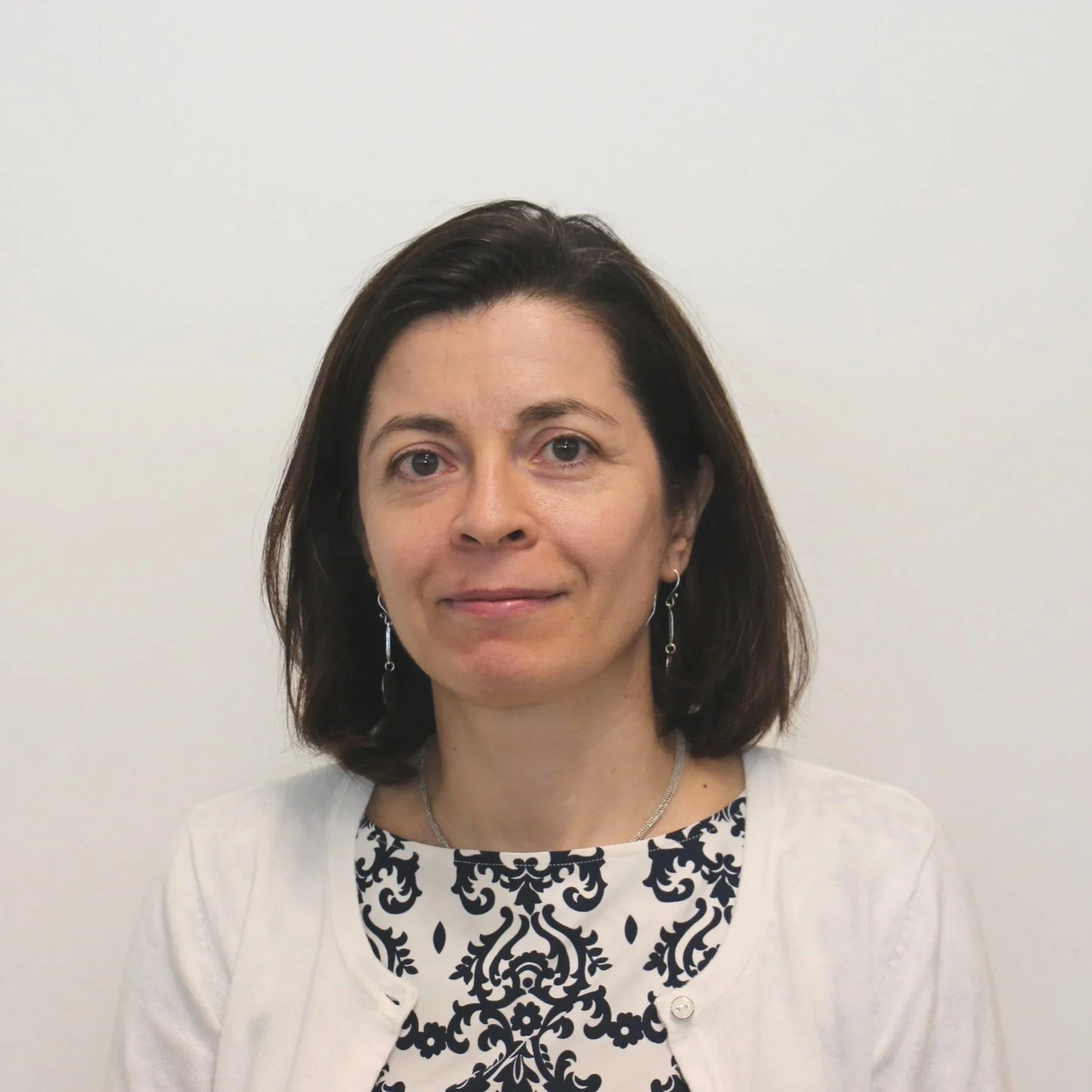 A woman with dark brown hair wearing earrings, a patterned black and white top, and a white cardigan standing against a plain white background.