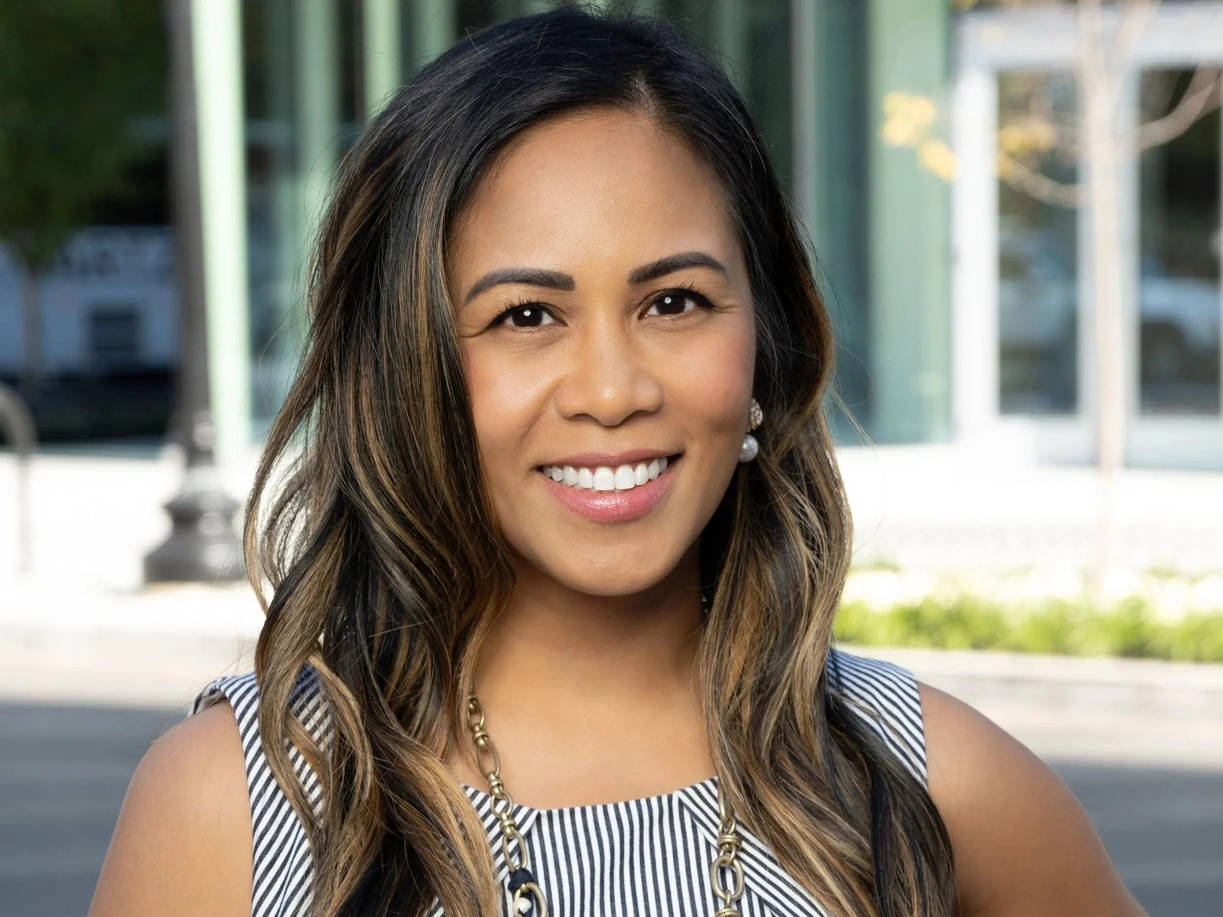 Portrait of a smiling woman with dark hair and light brown highlights, wearing a sleeveless striped top and a gold chain necklace, standing outdoors with glass building and greenery in the background.