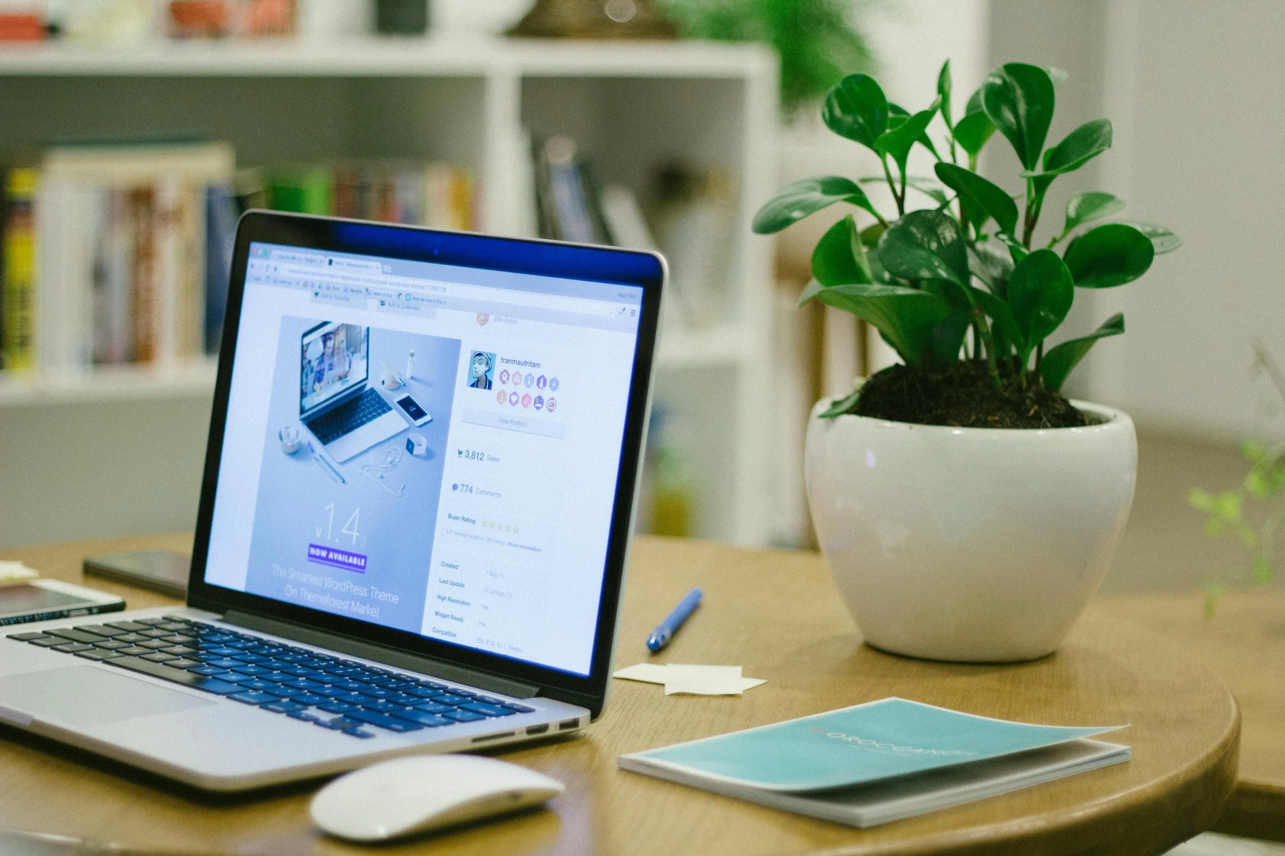 A laptop on a wooden desk with a bookshelf in the background, next to a white pot with a green plant, and some papers and a pen nearby.