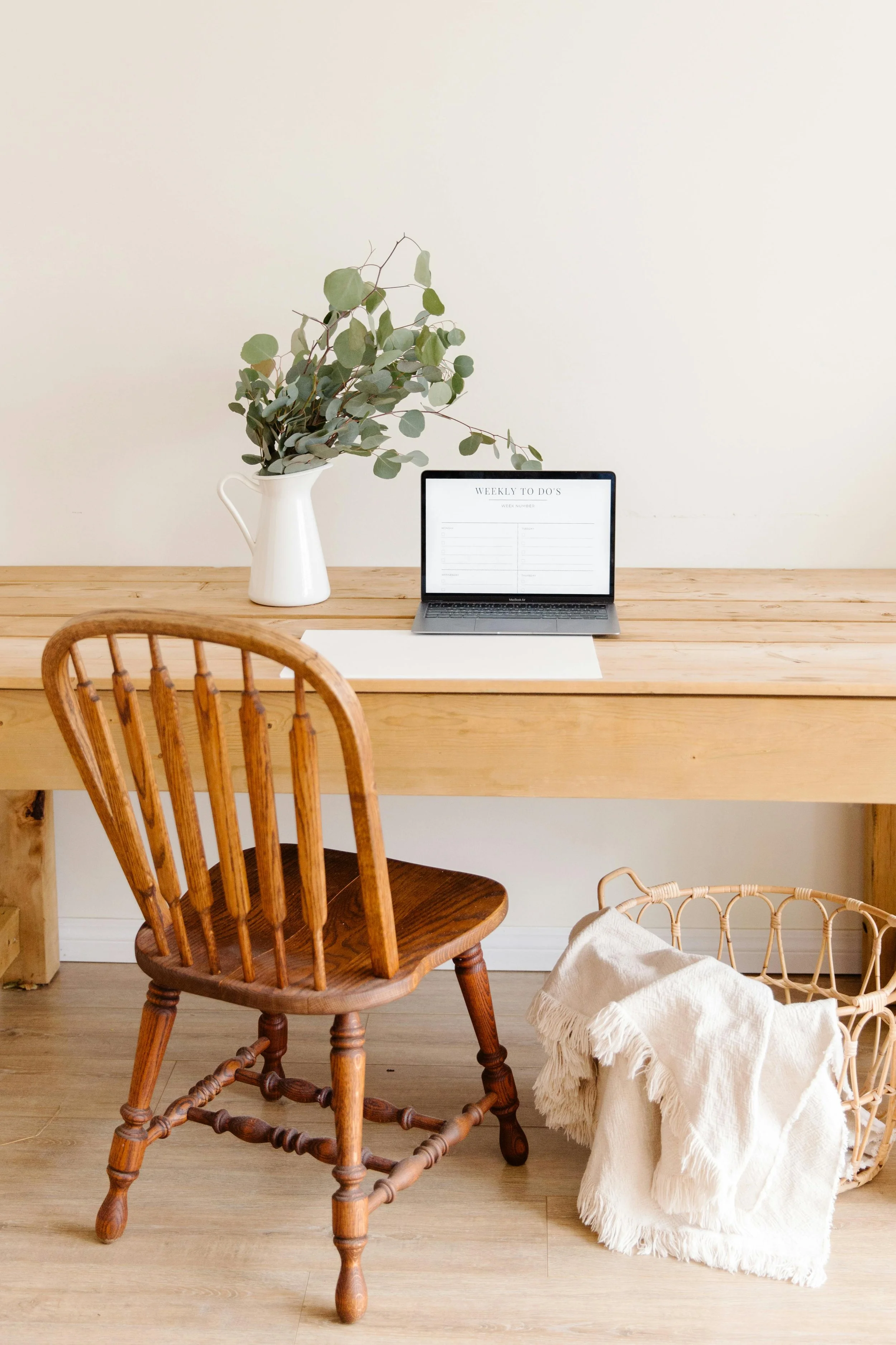 Minimalist home office with a wooden desk, a wooden chair, a laptop displaying a to-do list, a white vase with green eucalyptus leaves, and a woven basket with a blanket on the floor.
