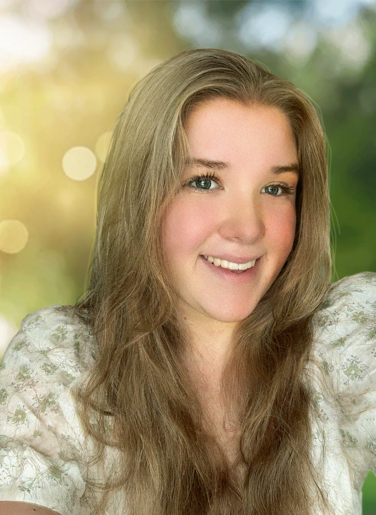 A woman with long, wavy blonde hair smiling at the camera, wearing a white floral blouse, with a blurred outdoor background of trees and sunlight.