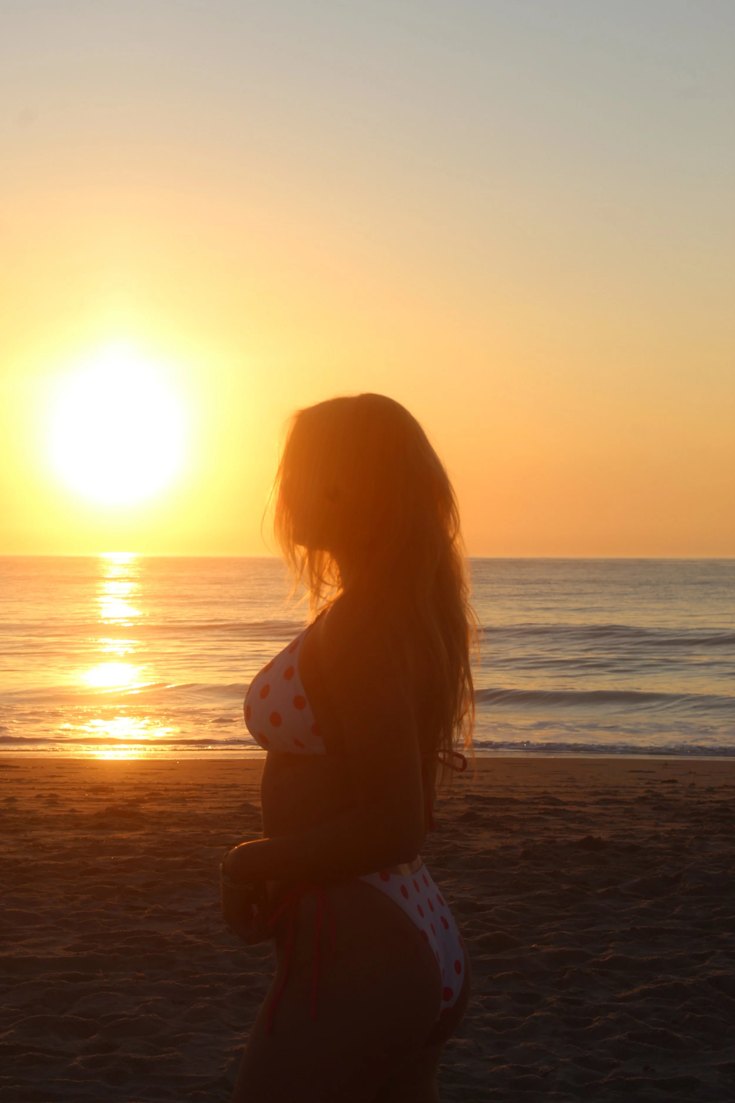 A woman in a polka dot bikini stands on the beach at sunset with her silhouette visible against the bright sun and the ocean.