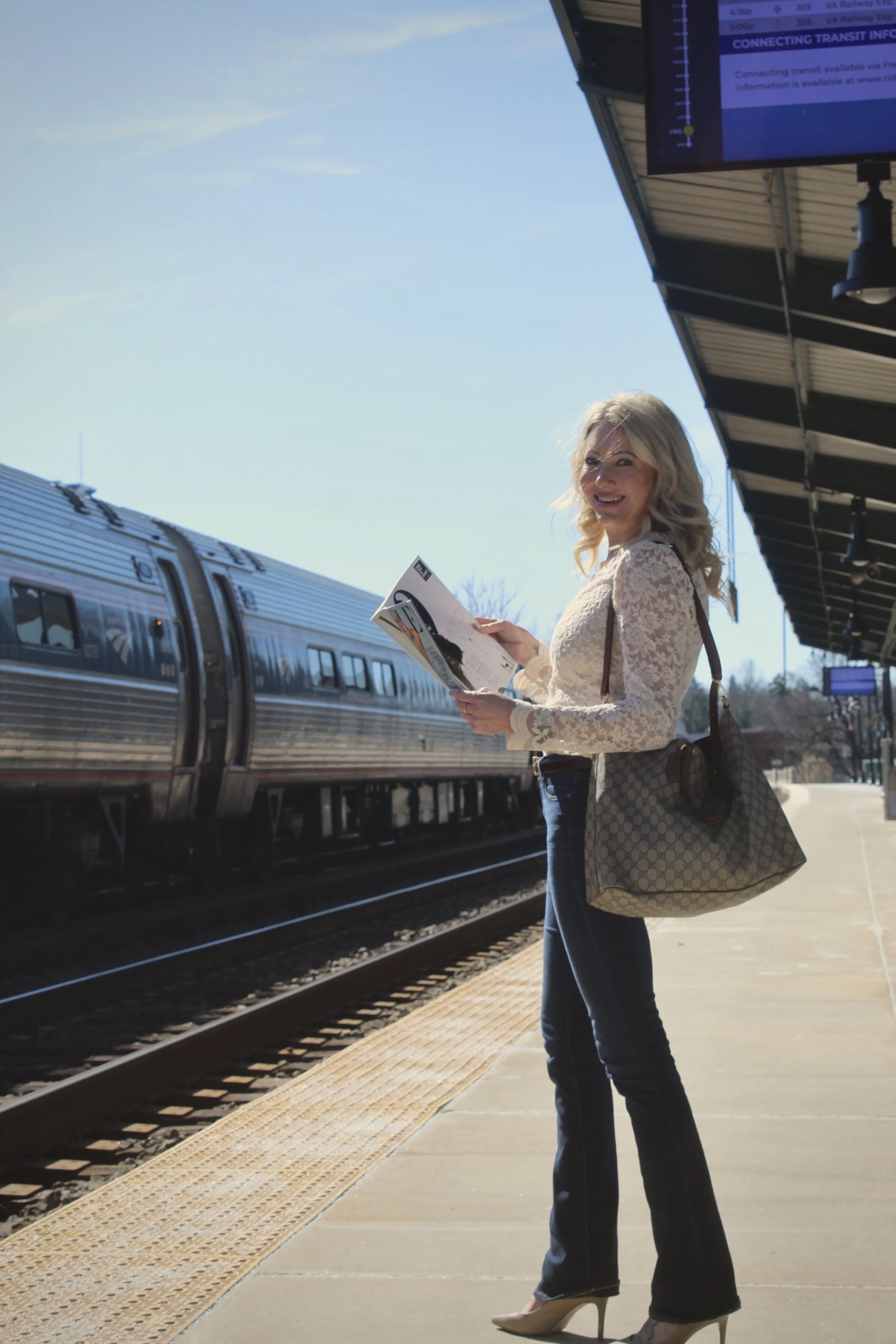 A woman with blonde hair standing on a train platform, looking at a pamphlet, with a silver train in the background on a sunny day.