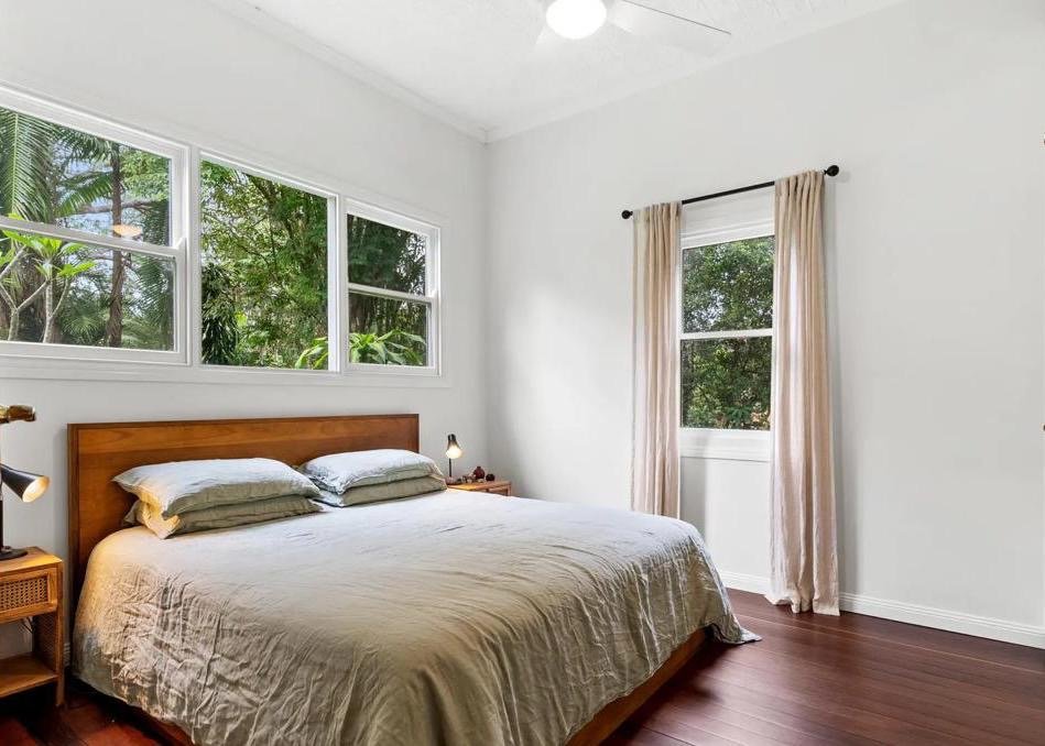 A bedroom with a large bed having a wooden headboard, beige bedding, and several pillows. There are two nightstands with small lamps, a large window with three sections and a view of green trees, and a door with beige curtains. The room has white walls and wooden floors.