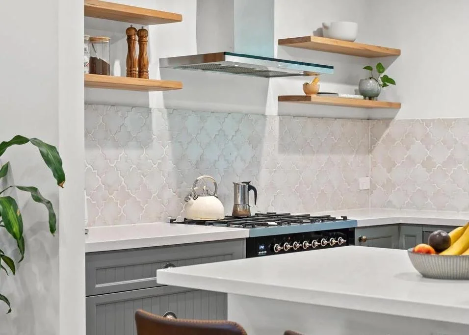 Kitchen with gray cabinets, white countertops, and open wooden shelves holding jars, a bowl, a plant, and a small sculpture. There is a stove with a kettle and a cooking pot, and a fruit bowl with bananas and other fruits on the white island.