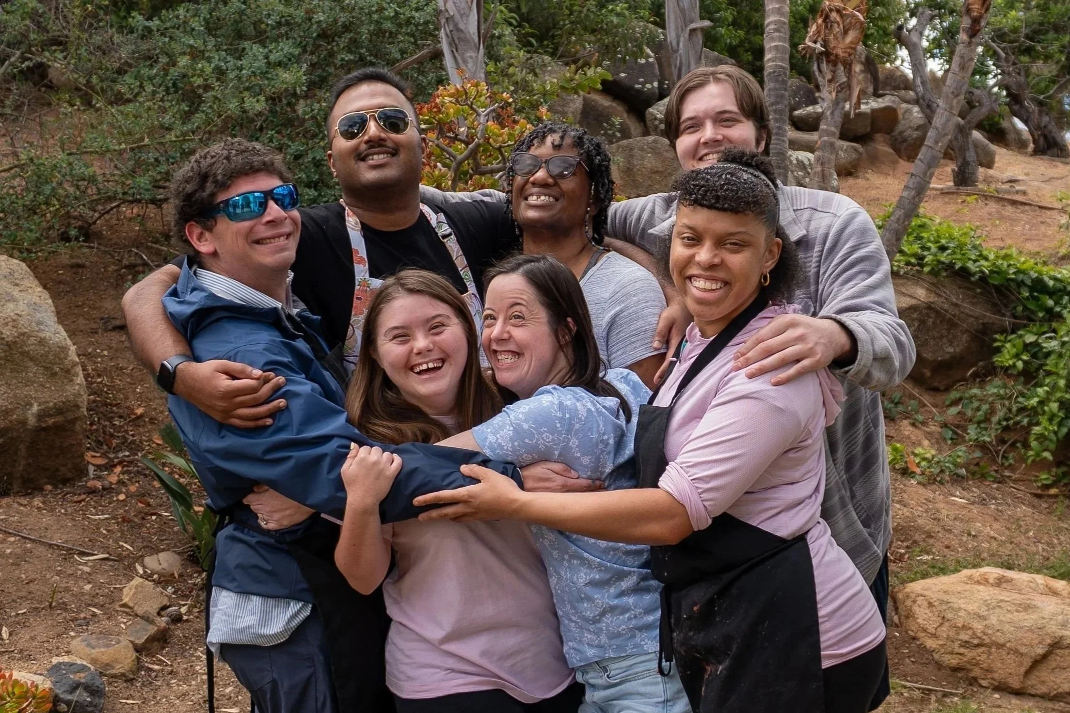 A diverse group of eight people, smiling and hugging in a circle outdoors, surrounded by rocks, trees, and plants.