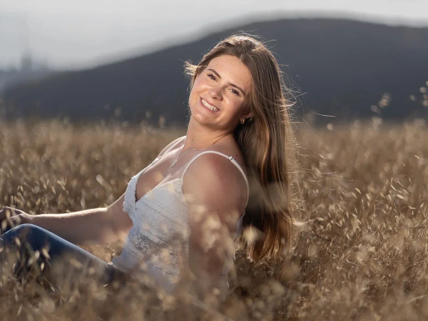 A young woman with long brown hair and a white top smiling and sitting in a golden wheat field with mountain in the background.