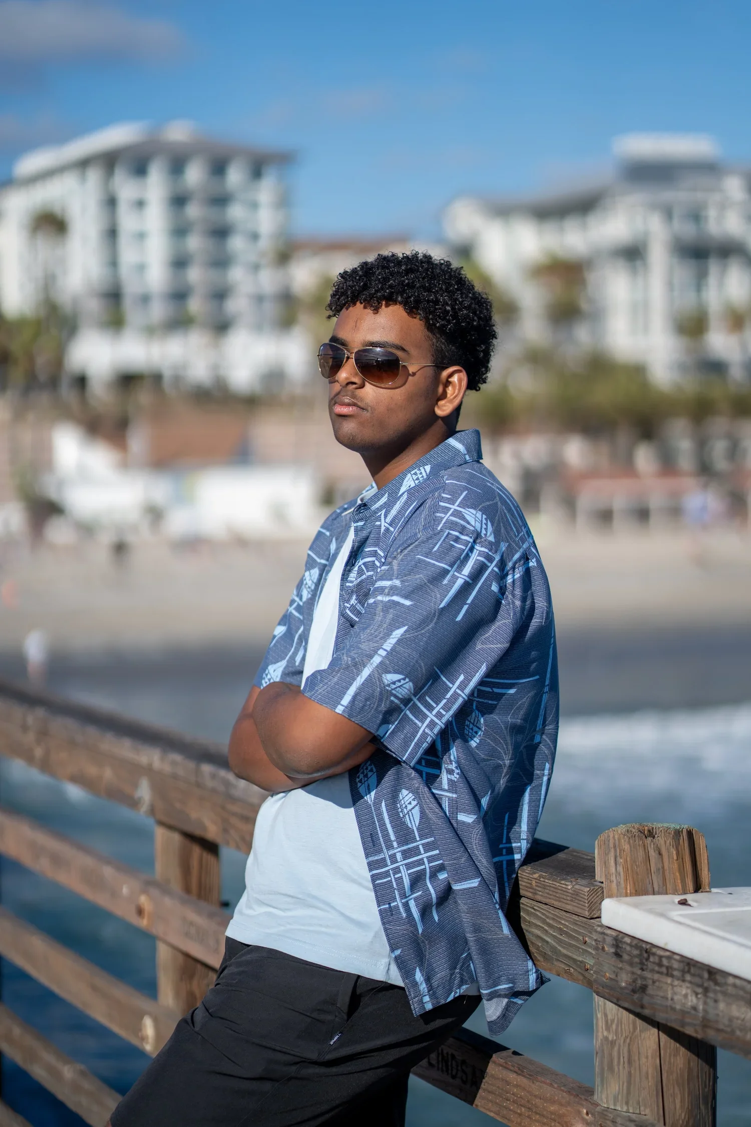 A young man with curly hair wearing sunglasses, a patterned blue shirt, and black shorts, standing with arms crossed on a wooden pier, with buildings and water in the background on a sunny day.
