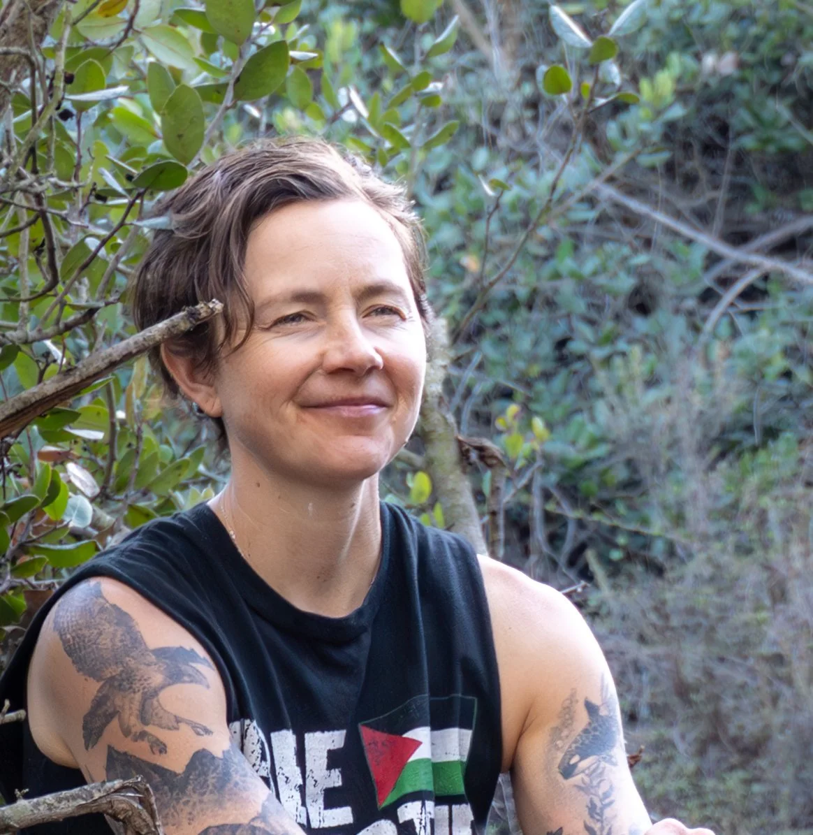 A woman with short brown hair and tattoos on her arms, smiling and sitting outside among green bushes.