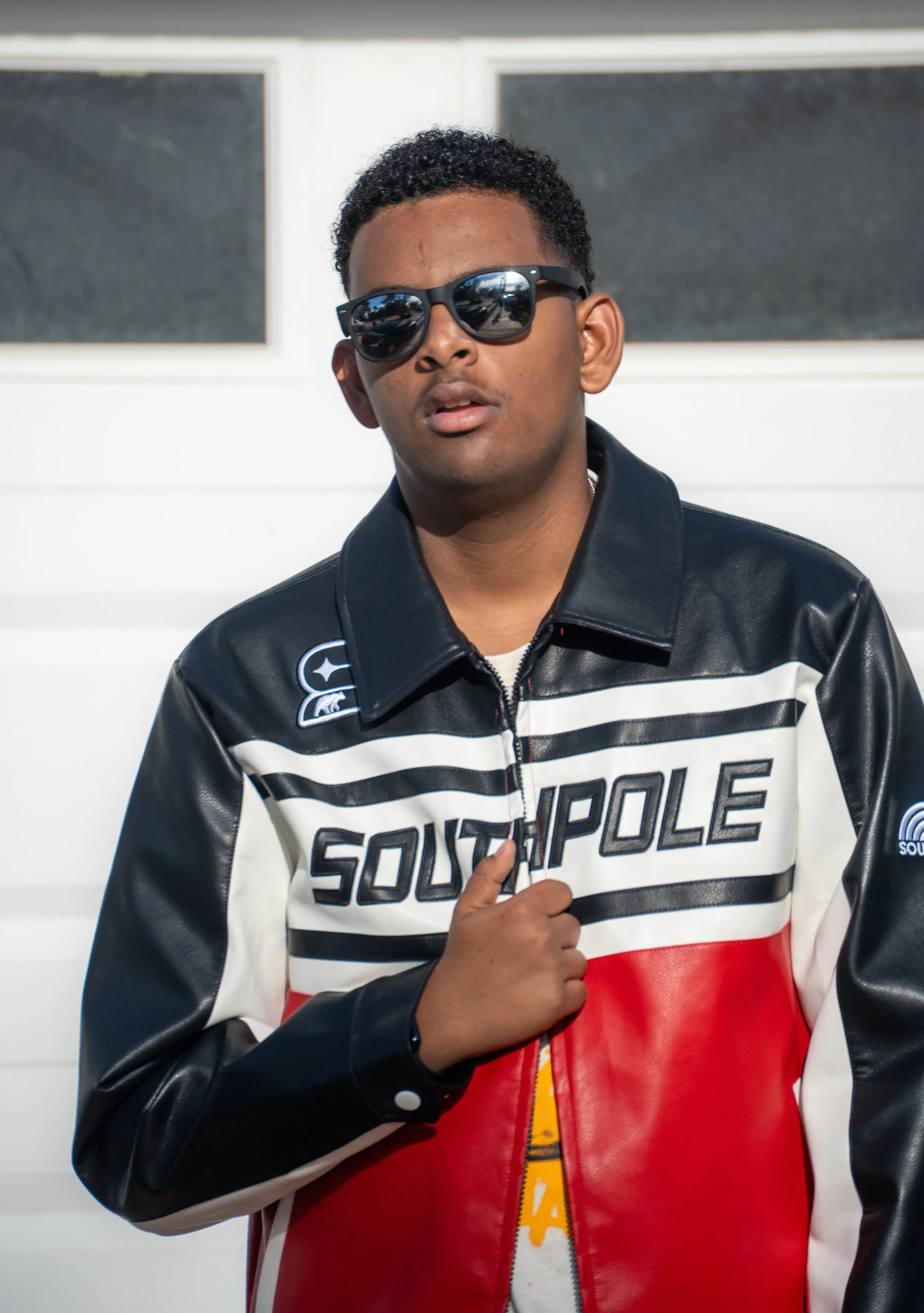 A young man wearing sunglasses and a color-blocked jacket with the word "Southpole" on it, standing outdoors in front of a white building with windows, holding the collar of his jacket.
